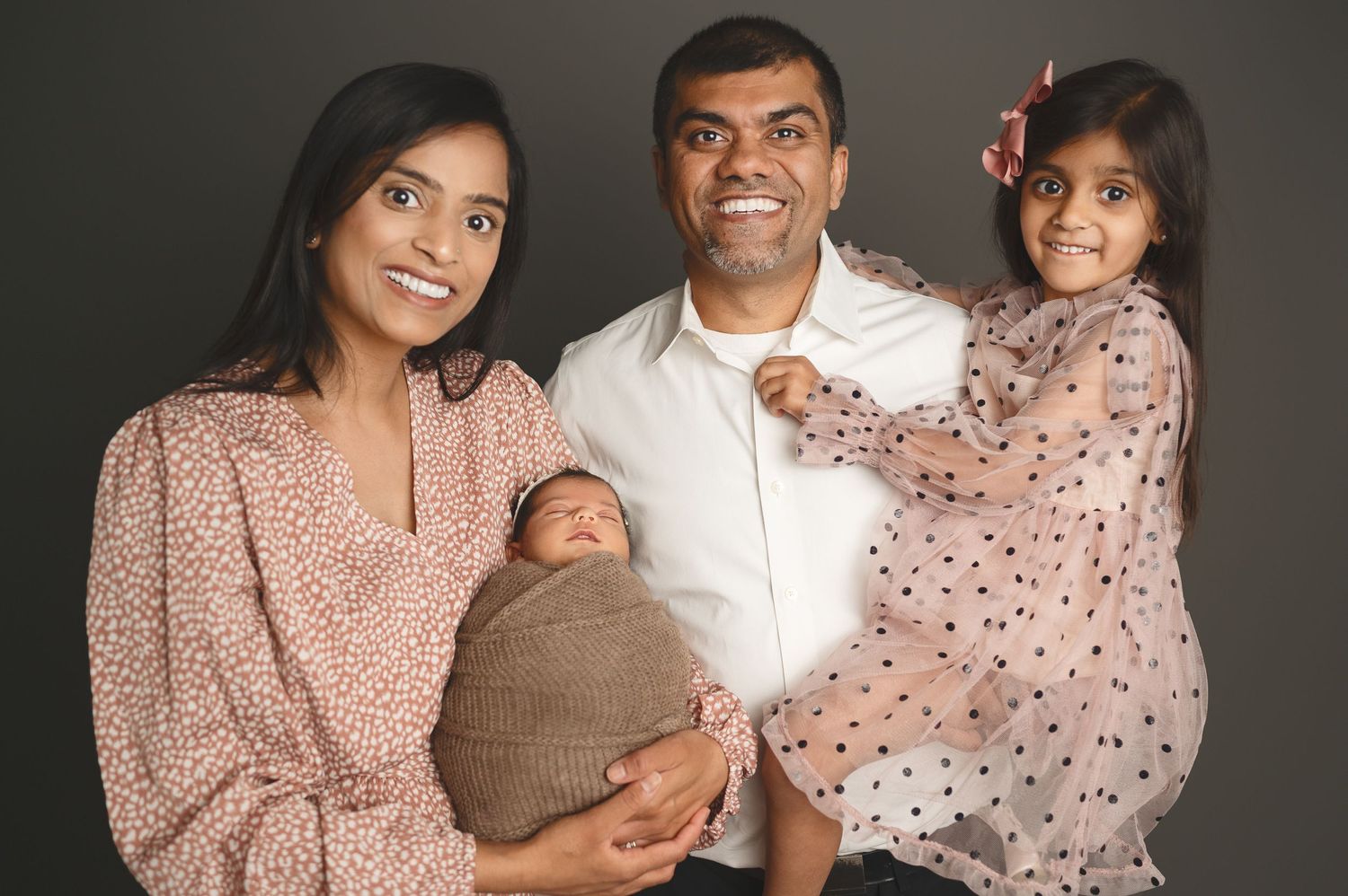 Family of four posing together in coordinating pink and white outfits against dark background.