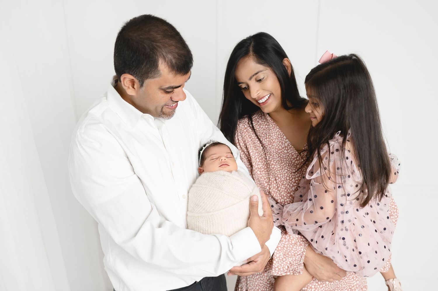 Family gathered around newborn baby in white studio setting wearing pink and white clothing.