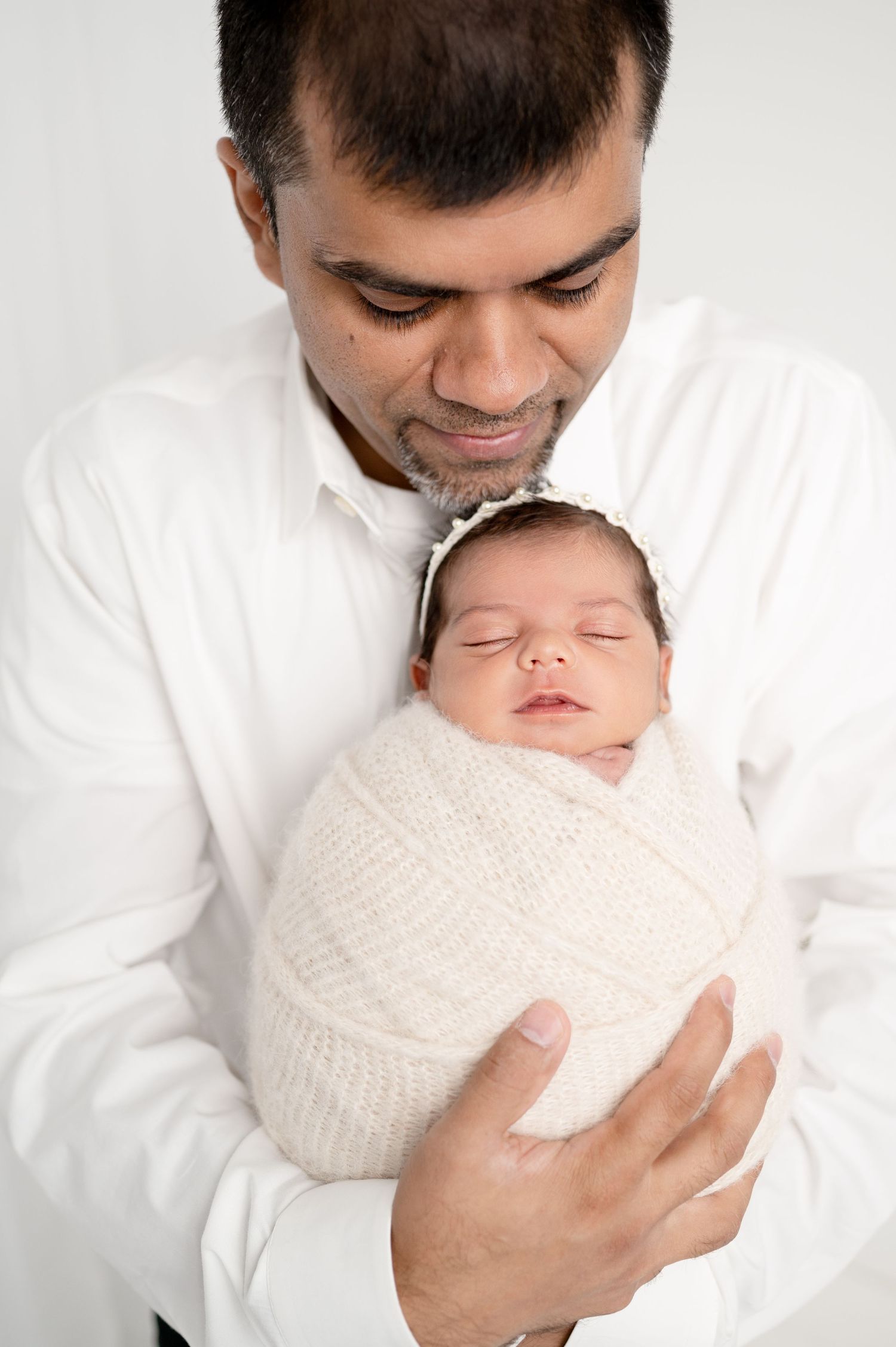 Tender moment between father in white shirt holding newborn wrapped in white against light background.