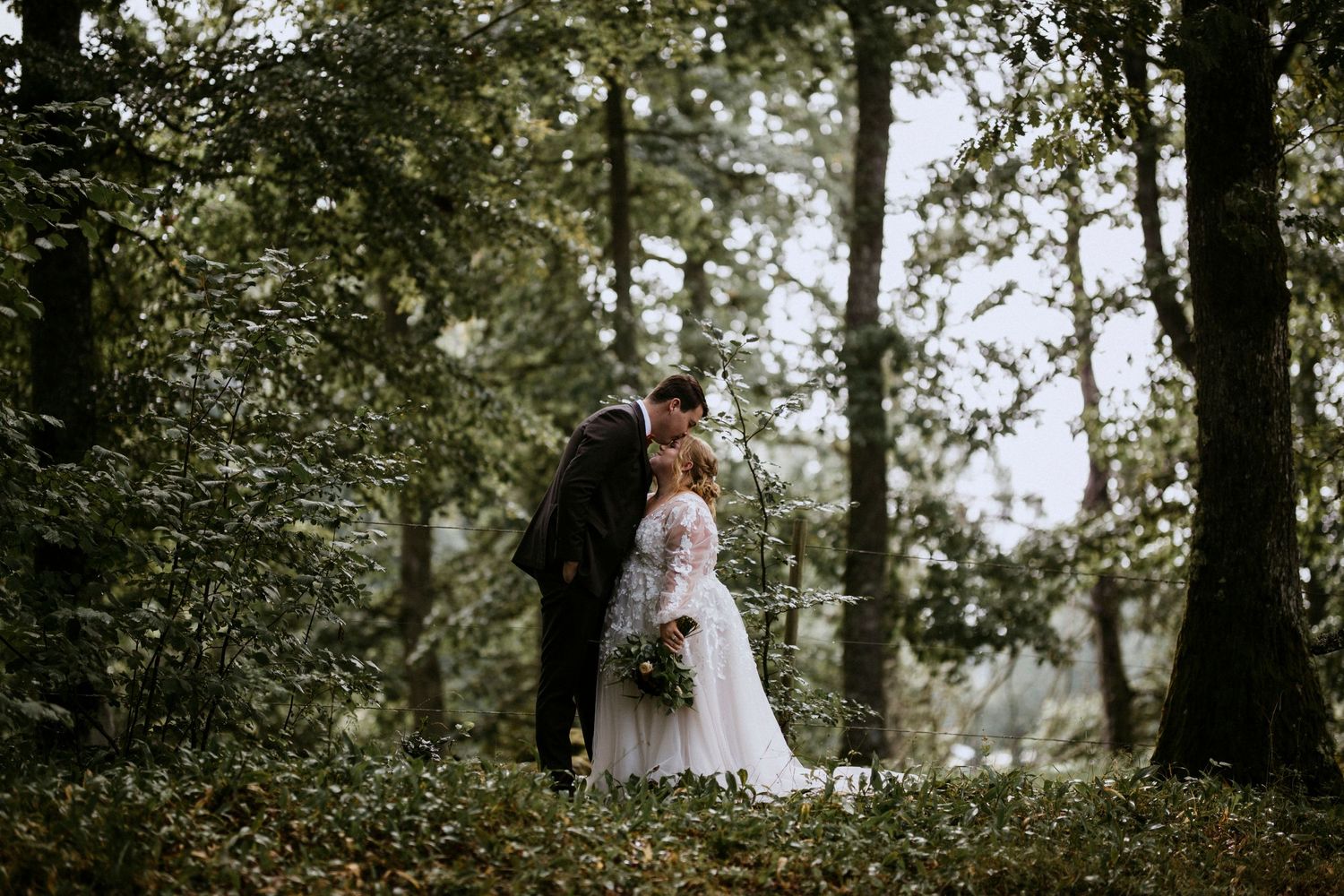 Romantic wedding couple embraces in a misty forest setting wearing formal attire.