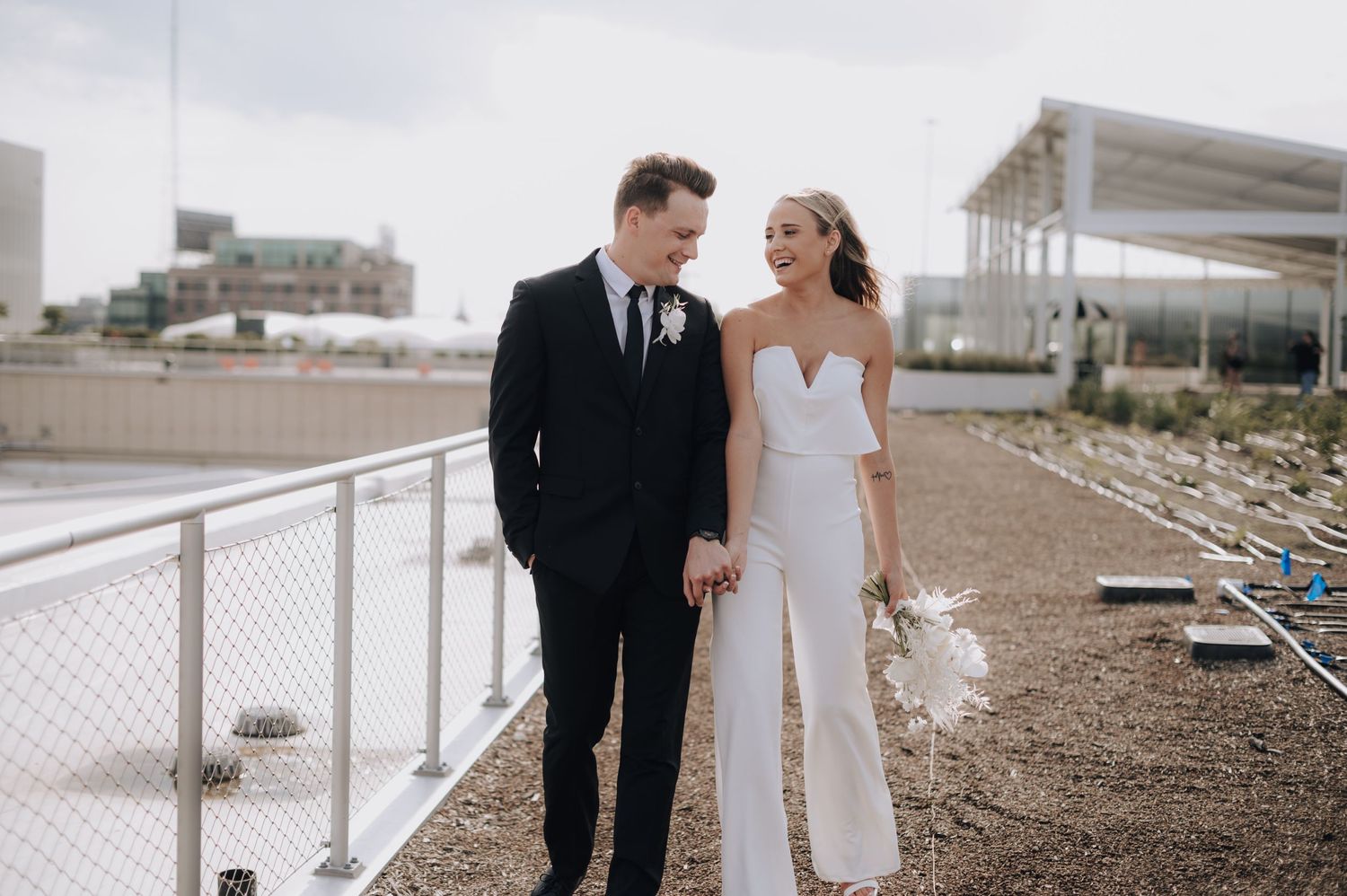 Elegant couple in wedding attire walks along modern waterfront railing.