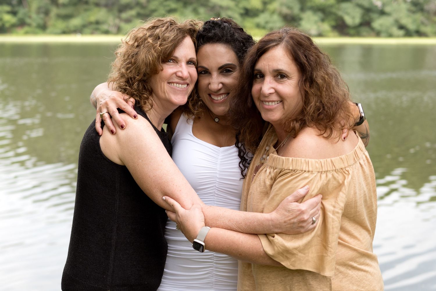 Three people hugging and laughing together by a serene lake.