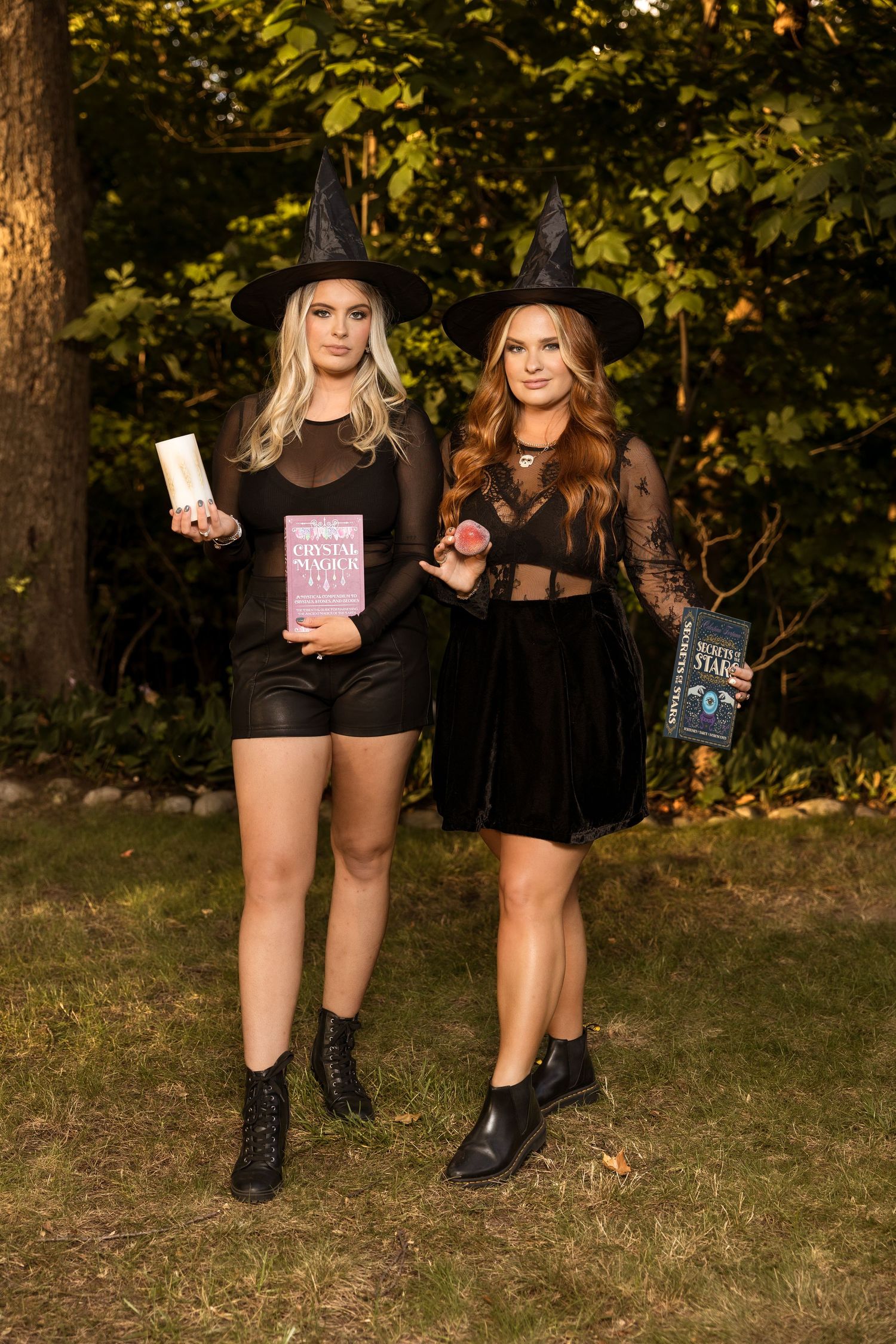 Two witches in black dresses and hats pose with spell books in an outdoor garden setting.