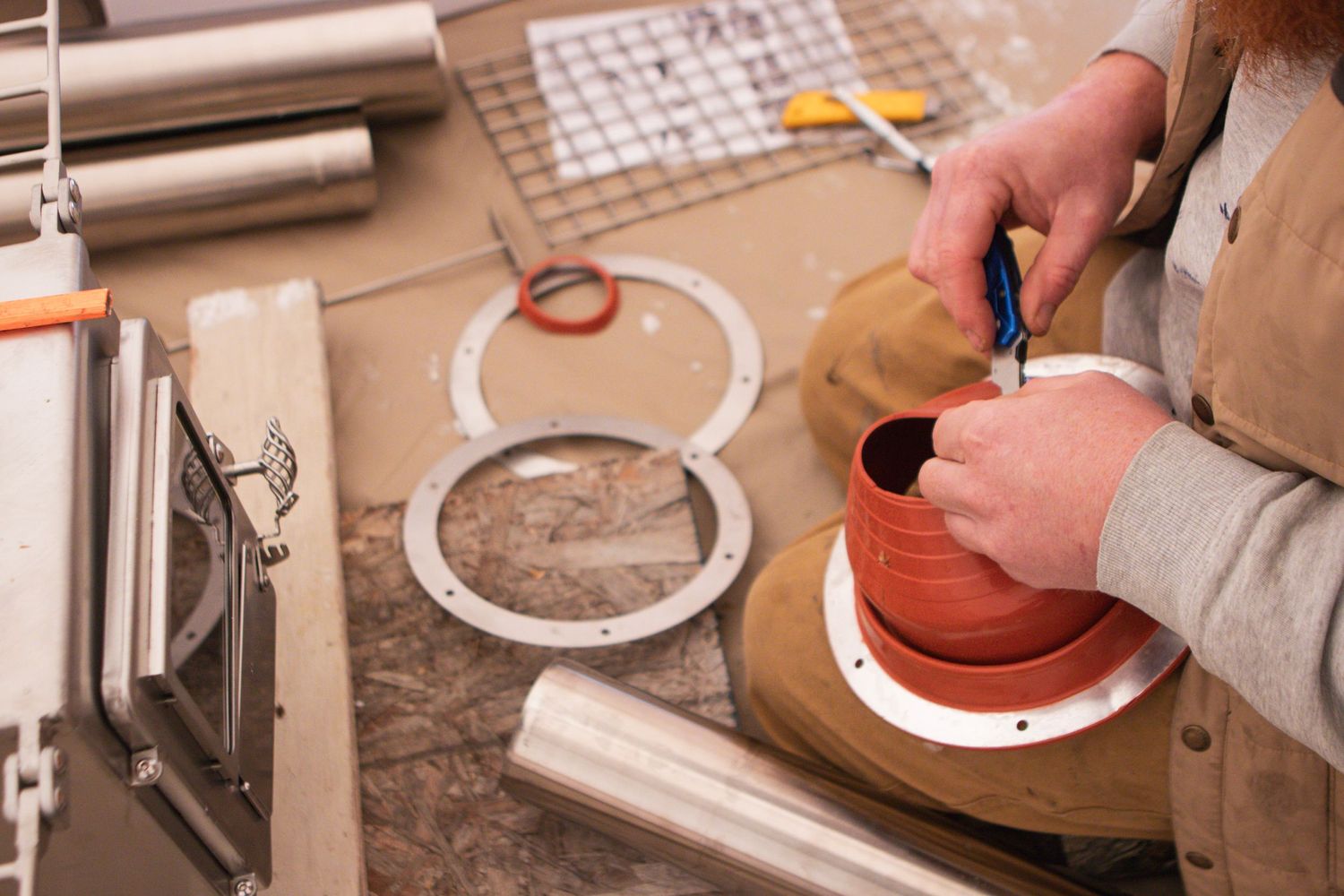 Hands assembling stove components on workbench with tools and parts.