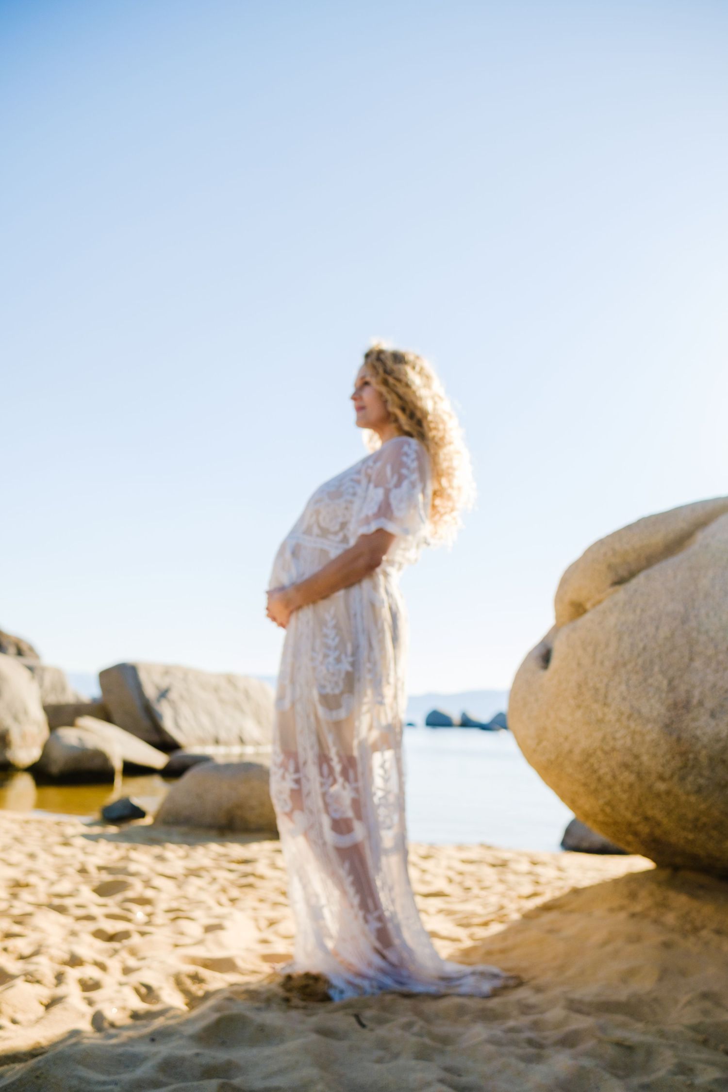 Ethereal maternity photo of a woman in a flowing white lace dress standing on a sandy beach among large boulders.