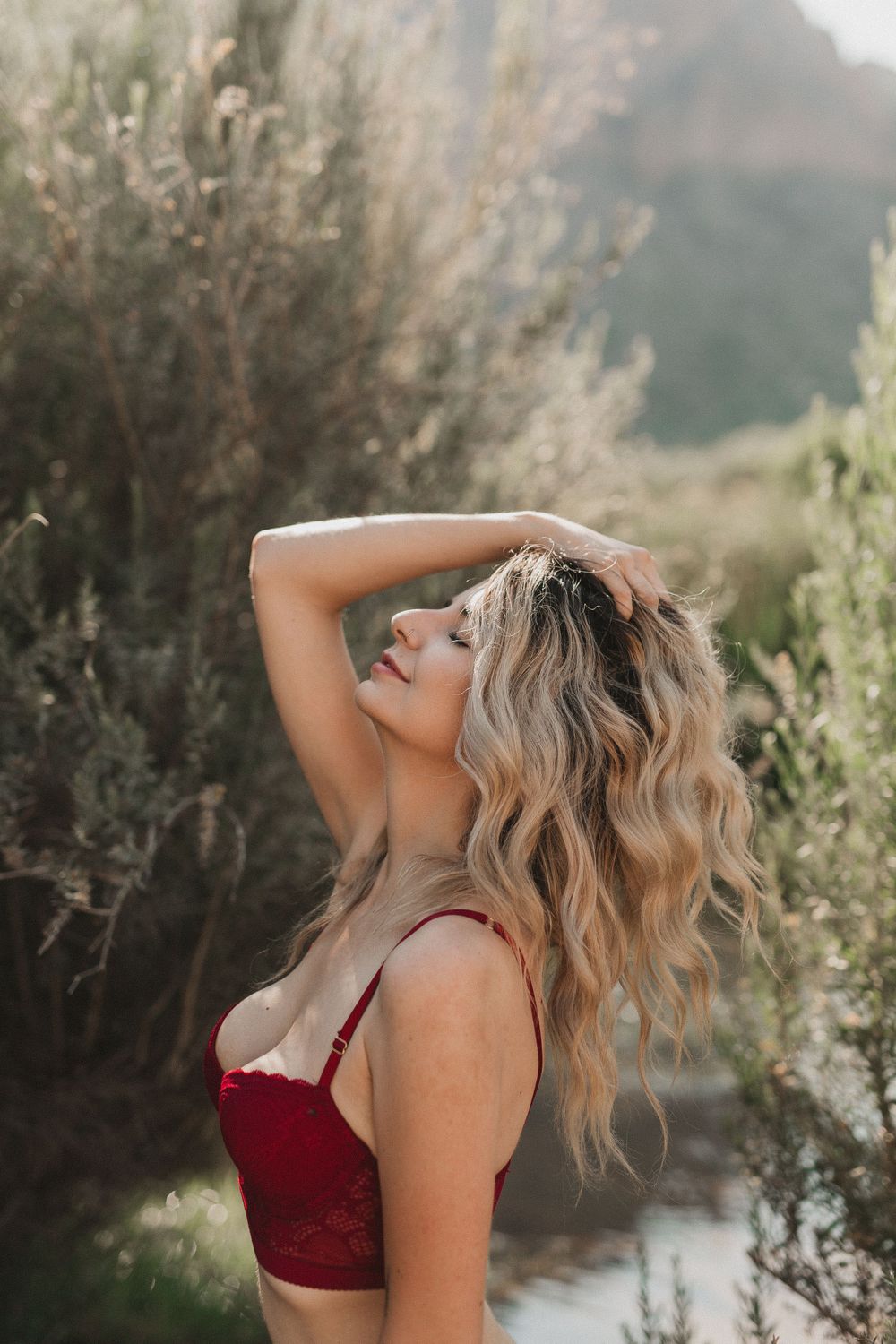 Blonde woman liberated in a river boudoir session, feeling free next to the water and lush plants wearing a red bra