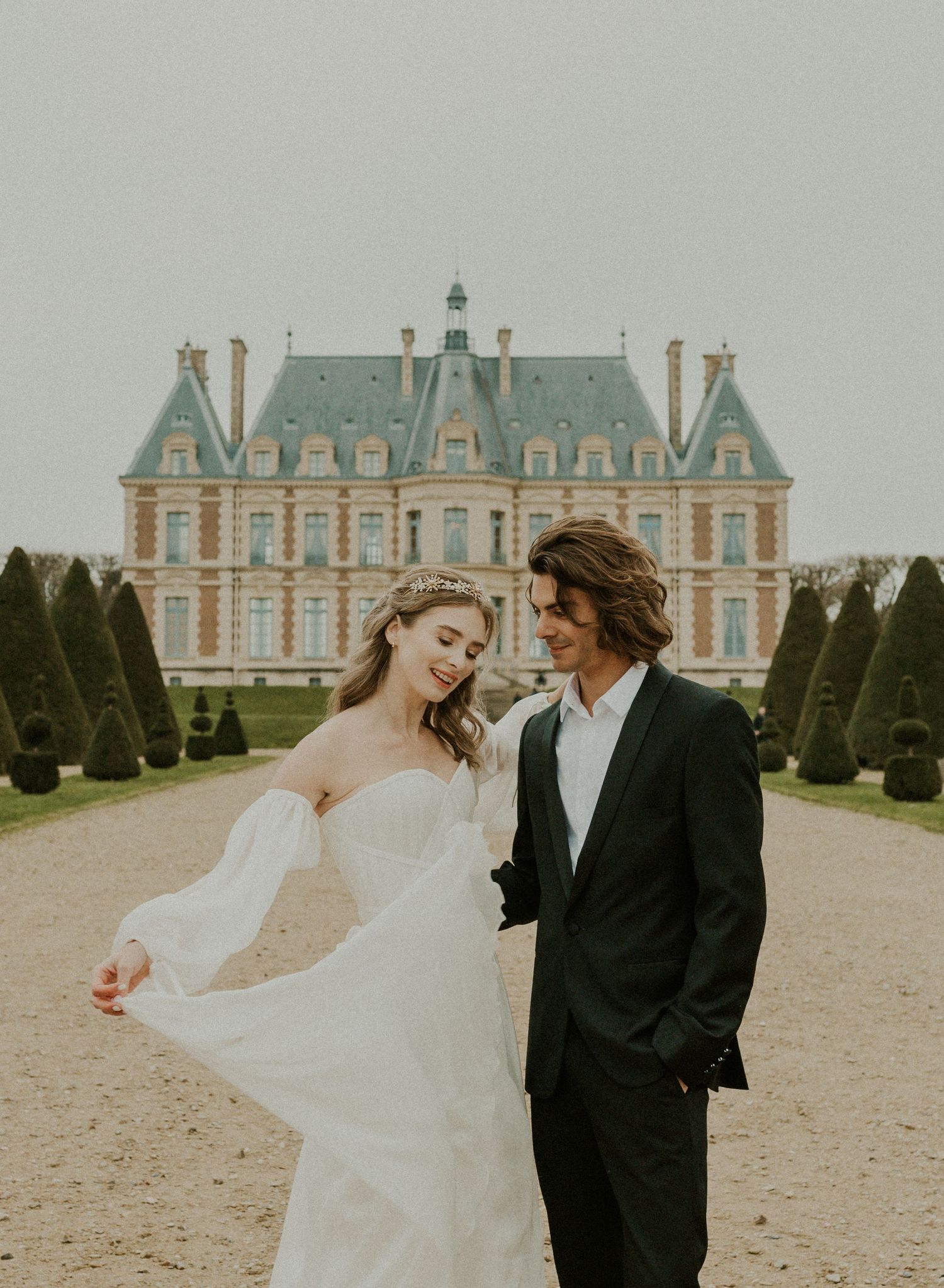 Romantic couple in wedding attire pose in front of grand French chateau with manicured gardens.