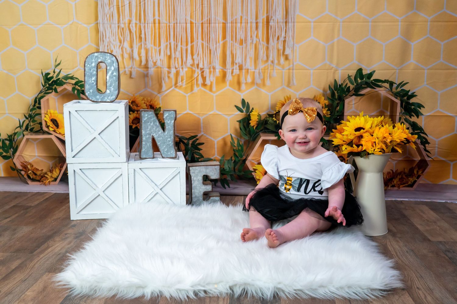 Happy baby girl celebrating first birthday sitting on white fur rug with sunflower decorations and ONE blocks.