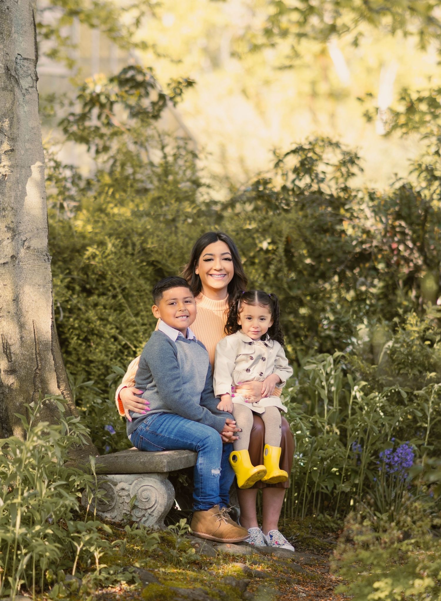 Family of 3 sitting on a bench next to a tree surrounded by beautiful foliage. Single mom of two. Young Kids