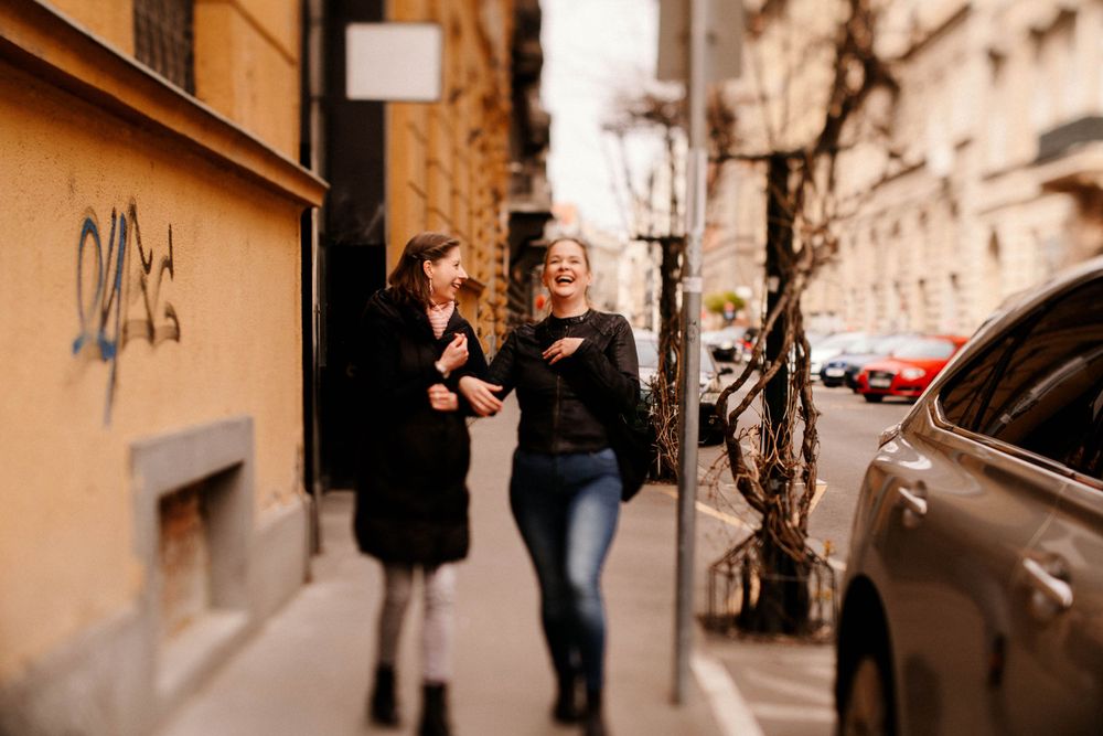 Friends walking and laughing together on urban sidewalk next to parked cars.