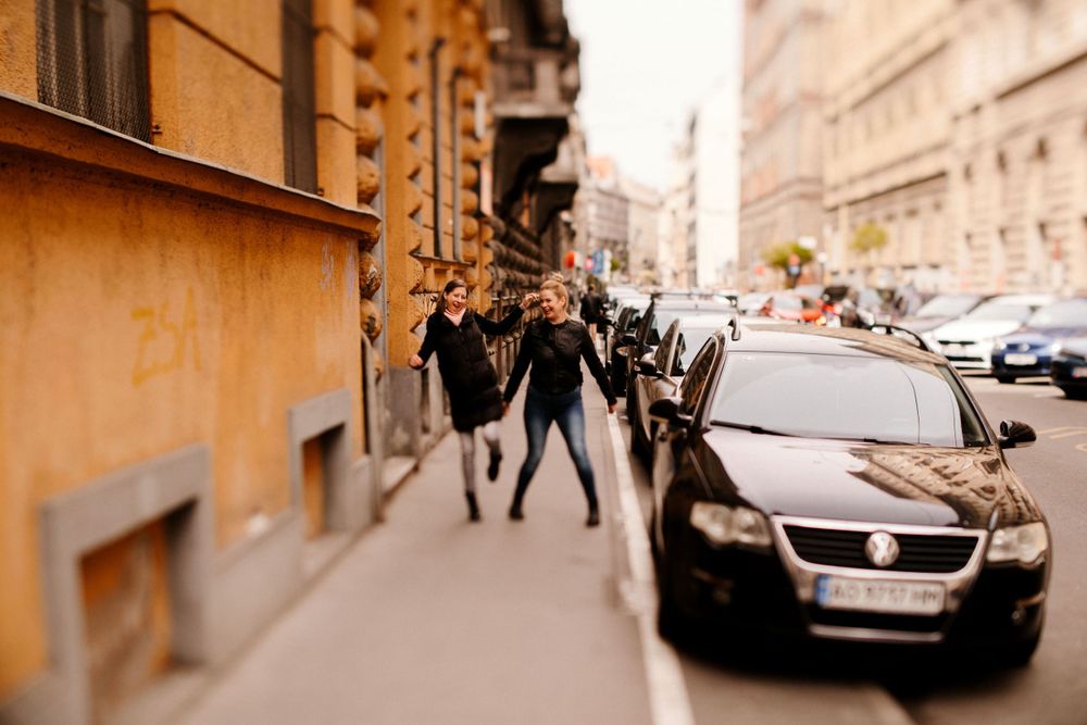 People walking on narrow European sidewalk with parked cars and historic buildings.