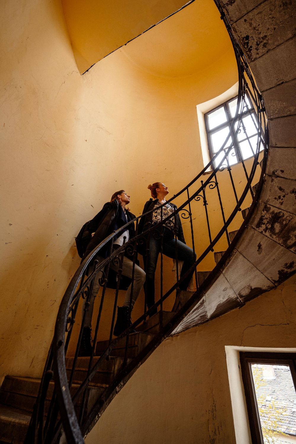 Two people on a spiral staircase with dramatic lighting through a window.
