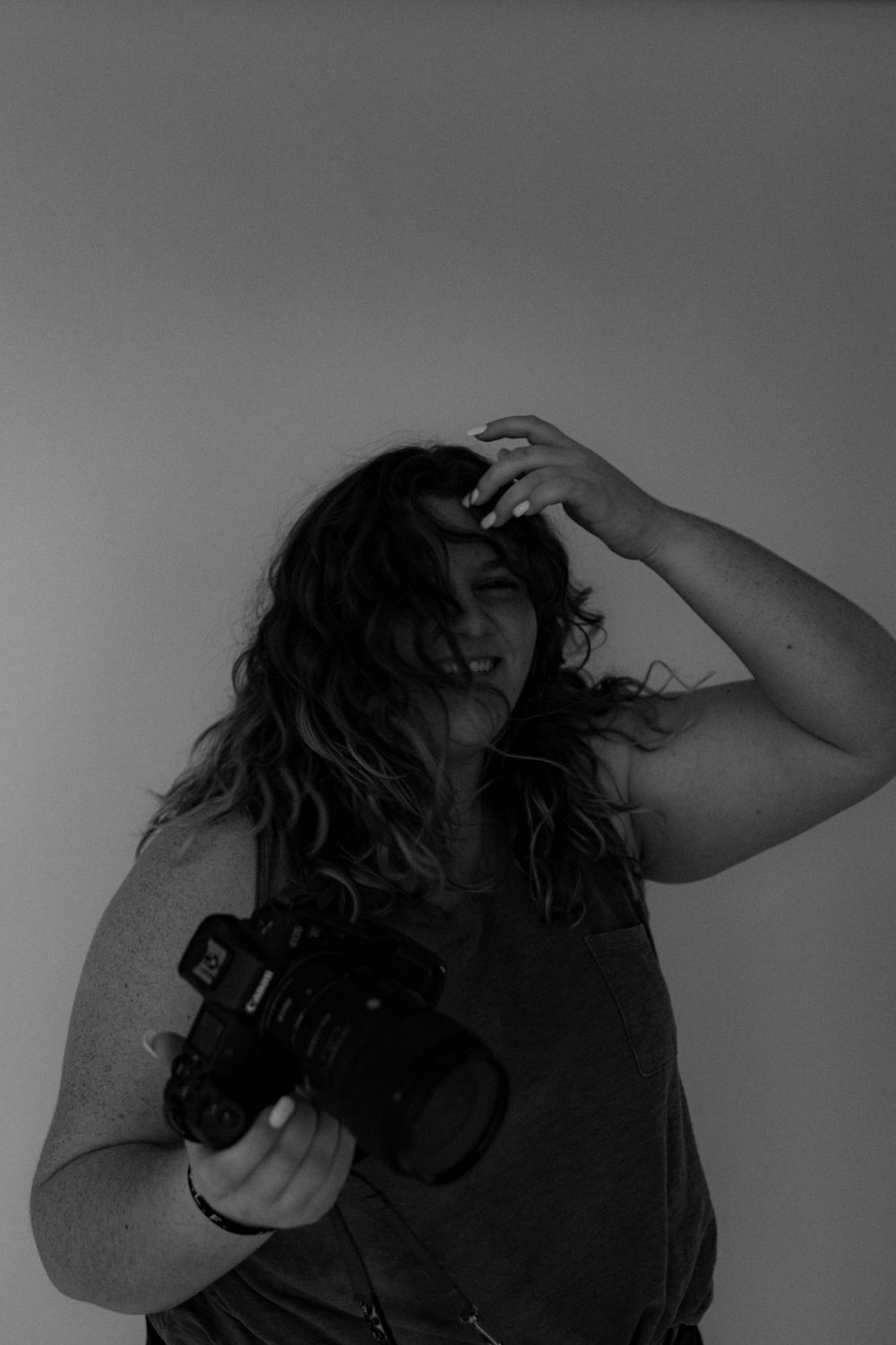 A photographer with curly hair holds a professional camera in a moody black and white portrait.