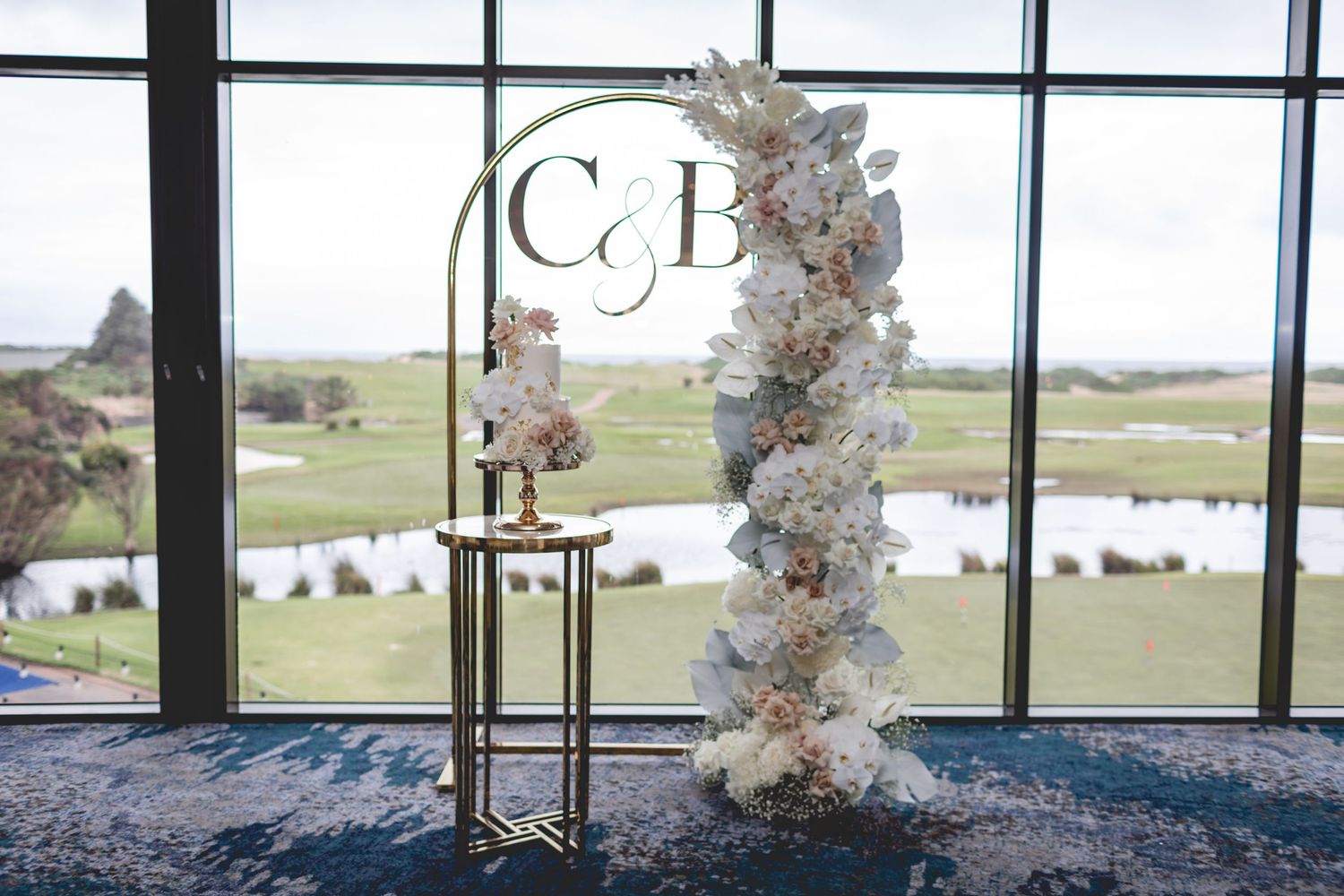 Elegant wedding monogram display with floral arch and gold stand against floor-to-ceiling windows overlooking scenic view.