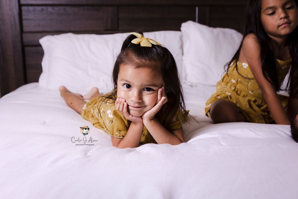 Young girl in yellow dress lying on white bedding with playful expression.