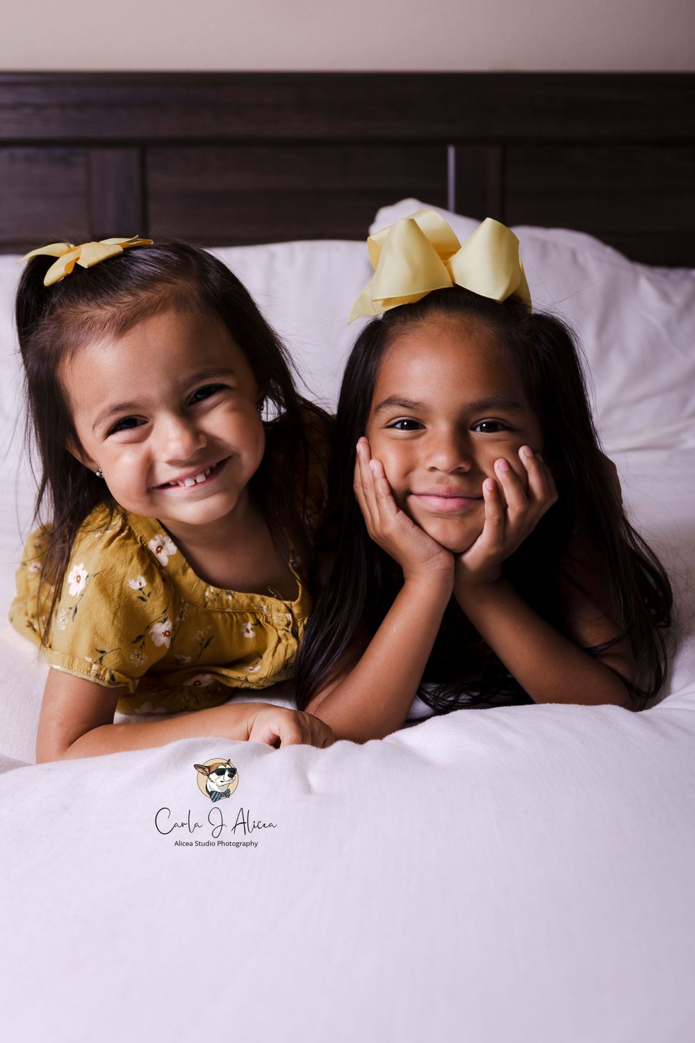 Two children in matching yellow dresses and bows pose together on white bedding.