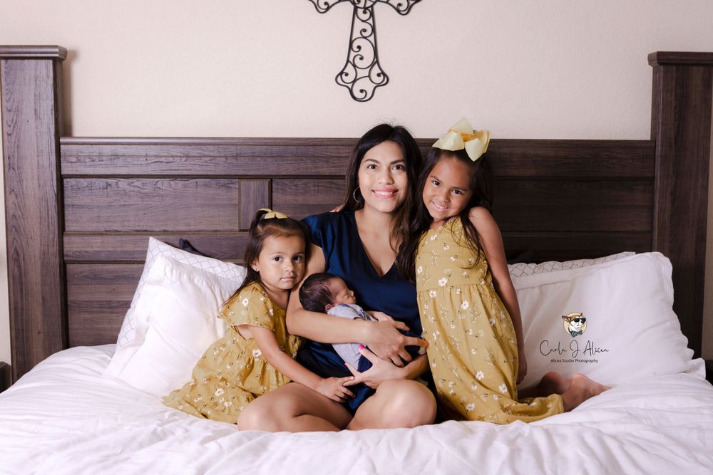 A tender family moment captured on a white bed with matching gold dresses and decorative cross on wall.