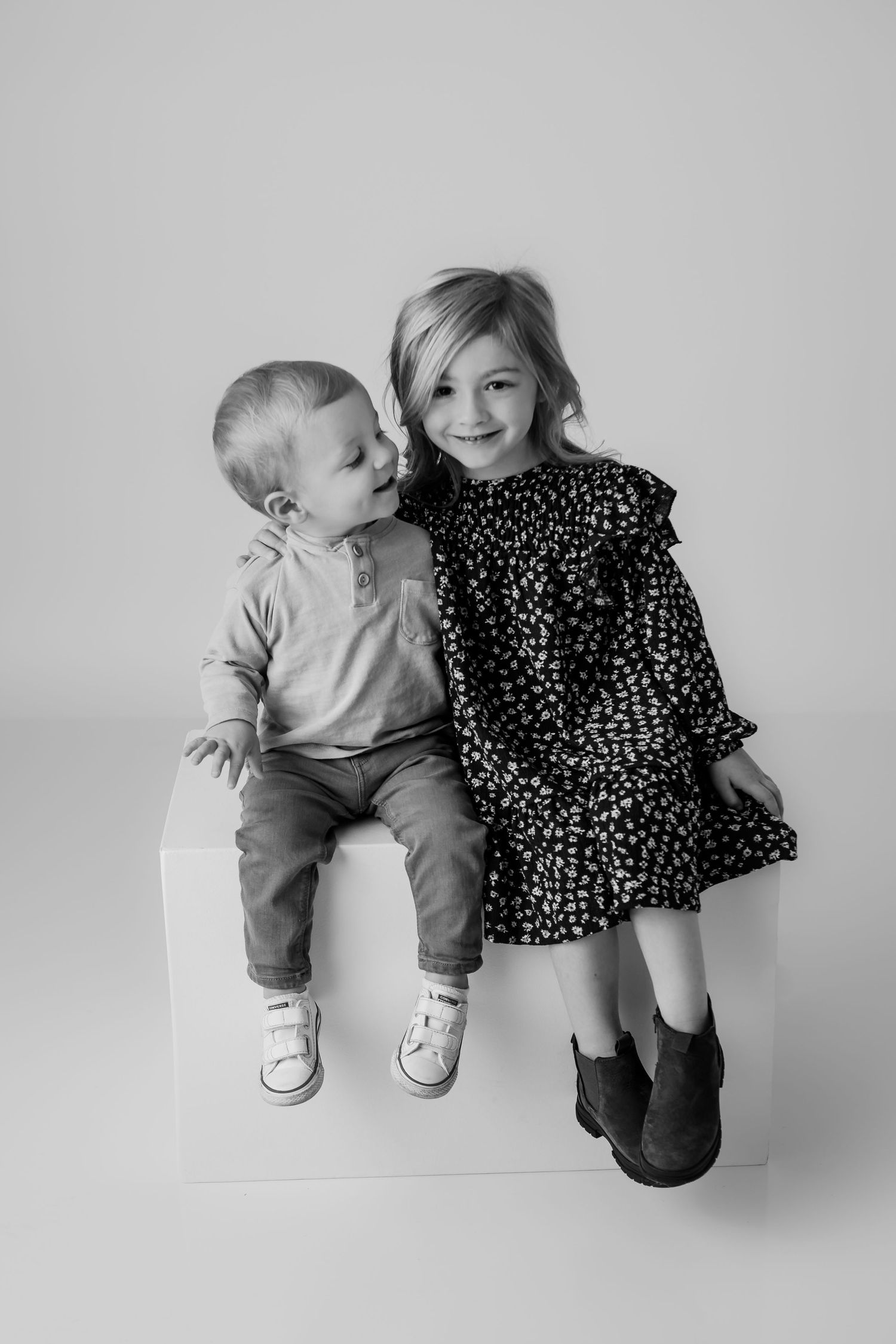 Black and white portrait of two children sitting together in front of a white backdrop to display their personalities in studio.