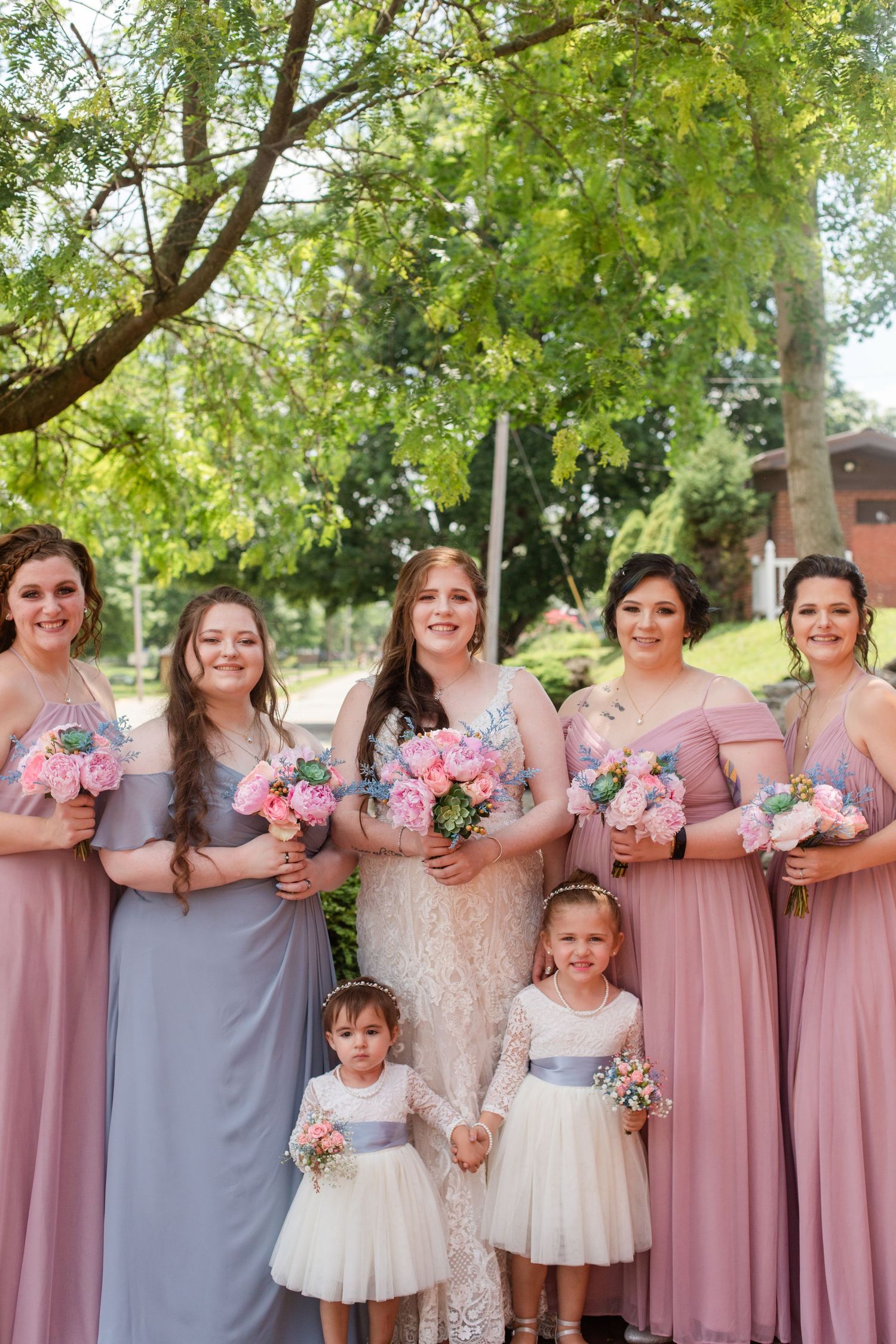 Wedding party with bridesmaids in pink dresses and flower girls in white poses under a leafy green tree.