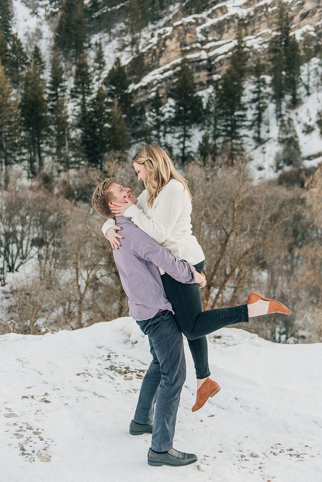 A romantic winter photoshoot in the snowy mountains with evergreen trees and rocky cliffs in the background.