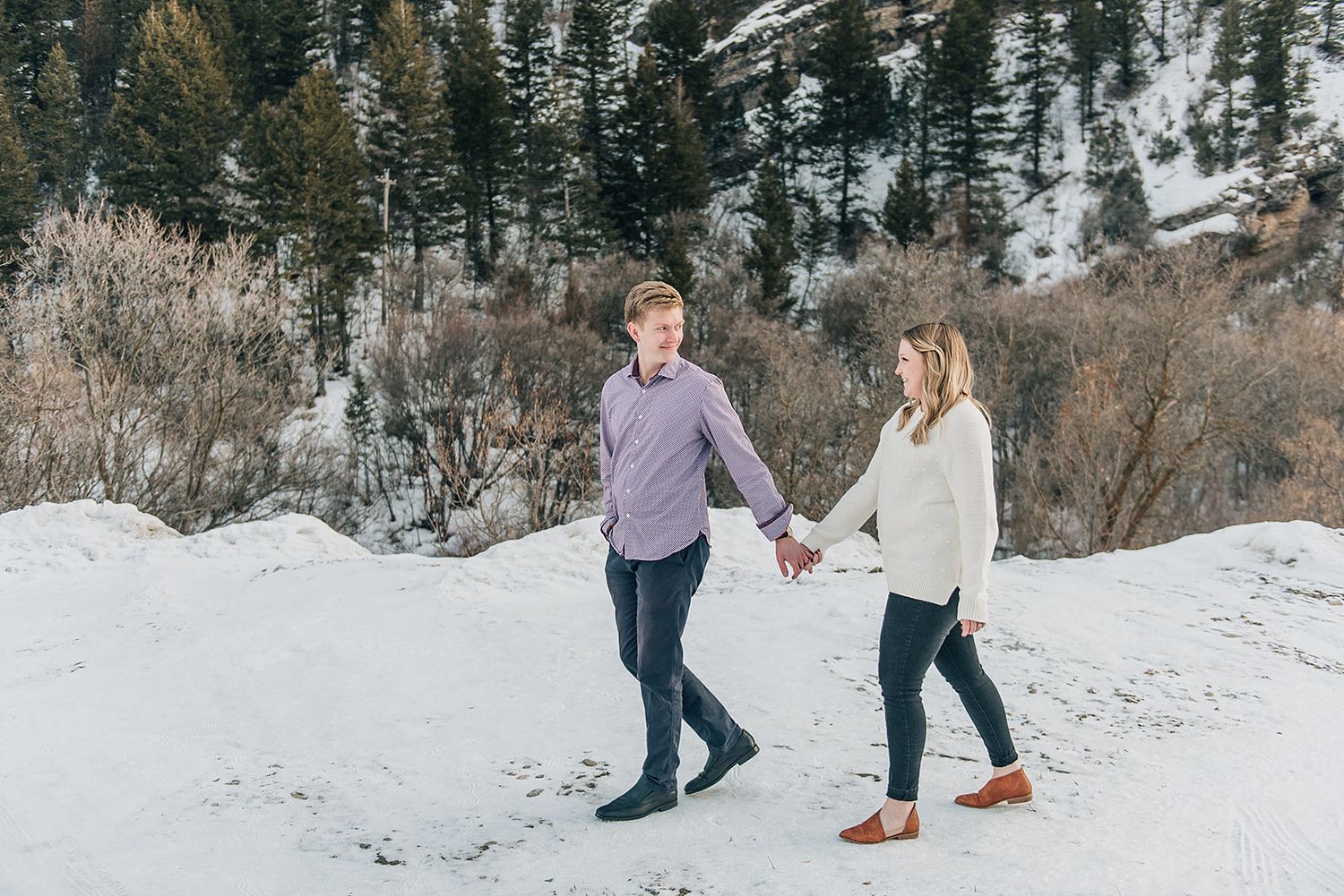 A winter engagement photo of a couple walking hand in hand in the snow with mountains behind them.