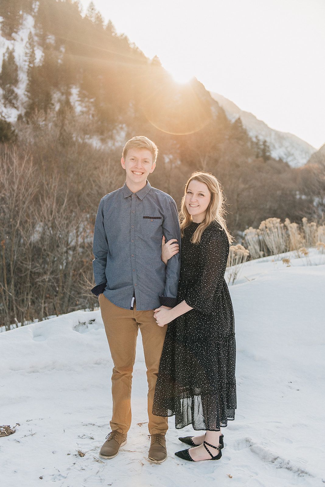 A couple poses together in front of snowy mountains during golden hour sunset.