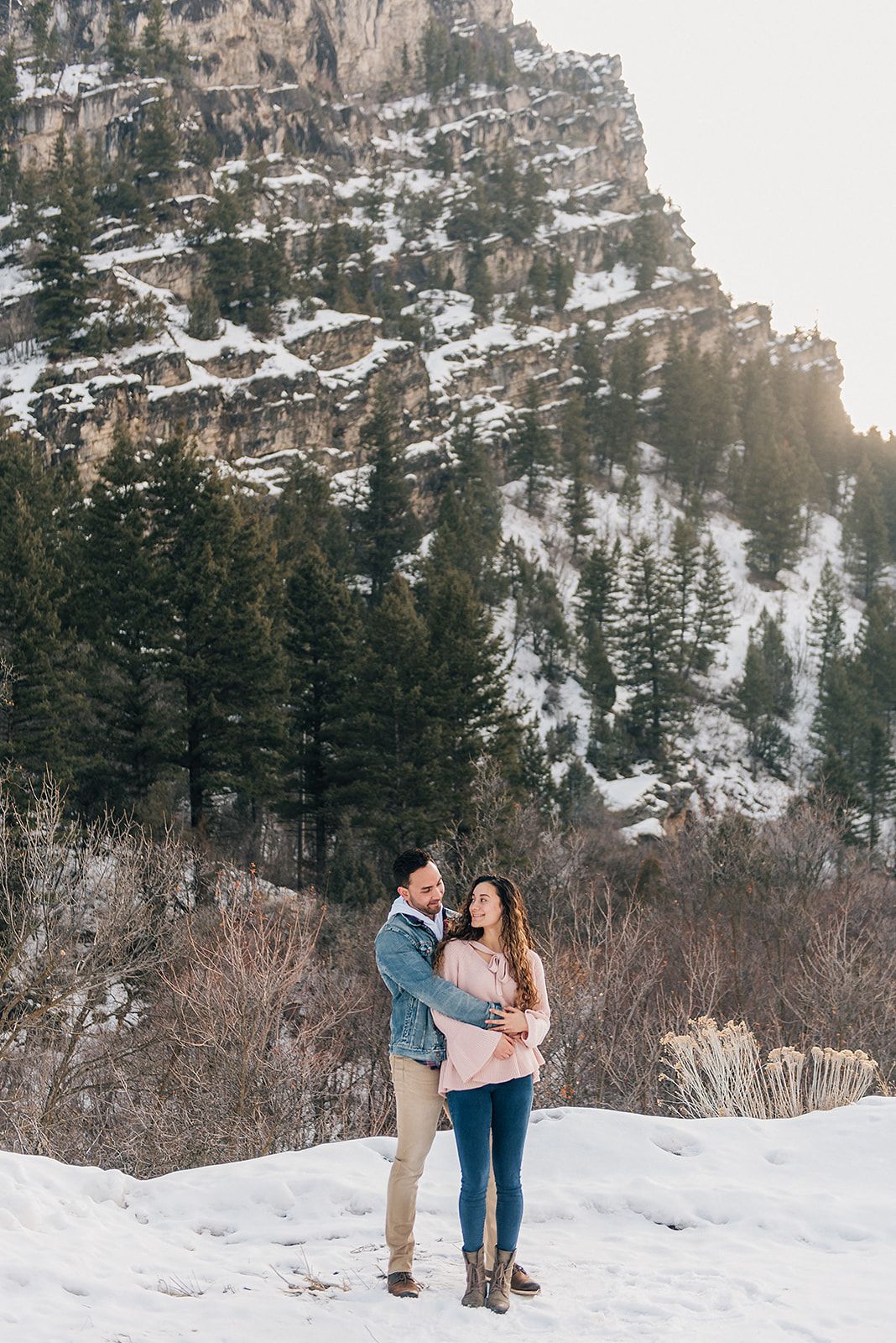 A couple poses together on a snowy mountain trail surrounded by evergreen trees and cliffs.