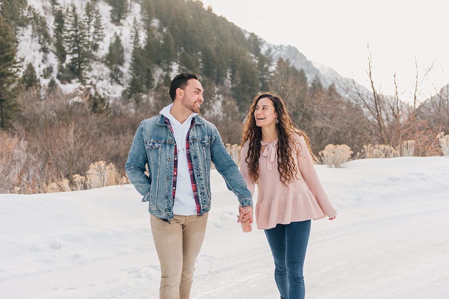 A couple walks hand in hand on a snowy path with mountains and pine trees in the background.