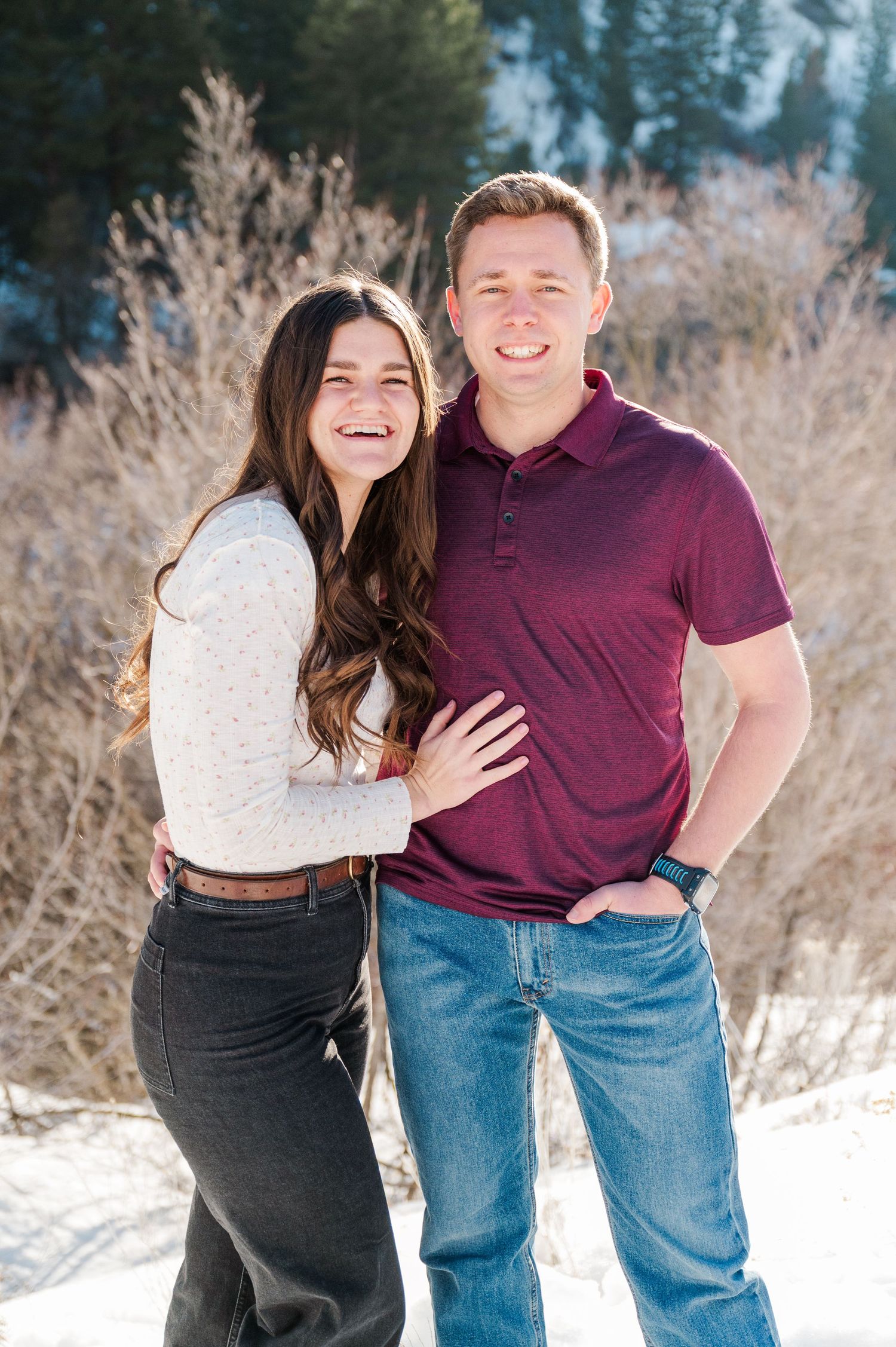 A couple poses together in winter attire with snow-capped mountains and bare branches behind them.
