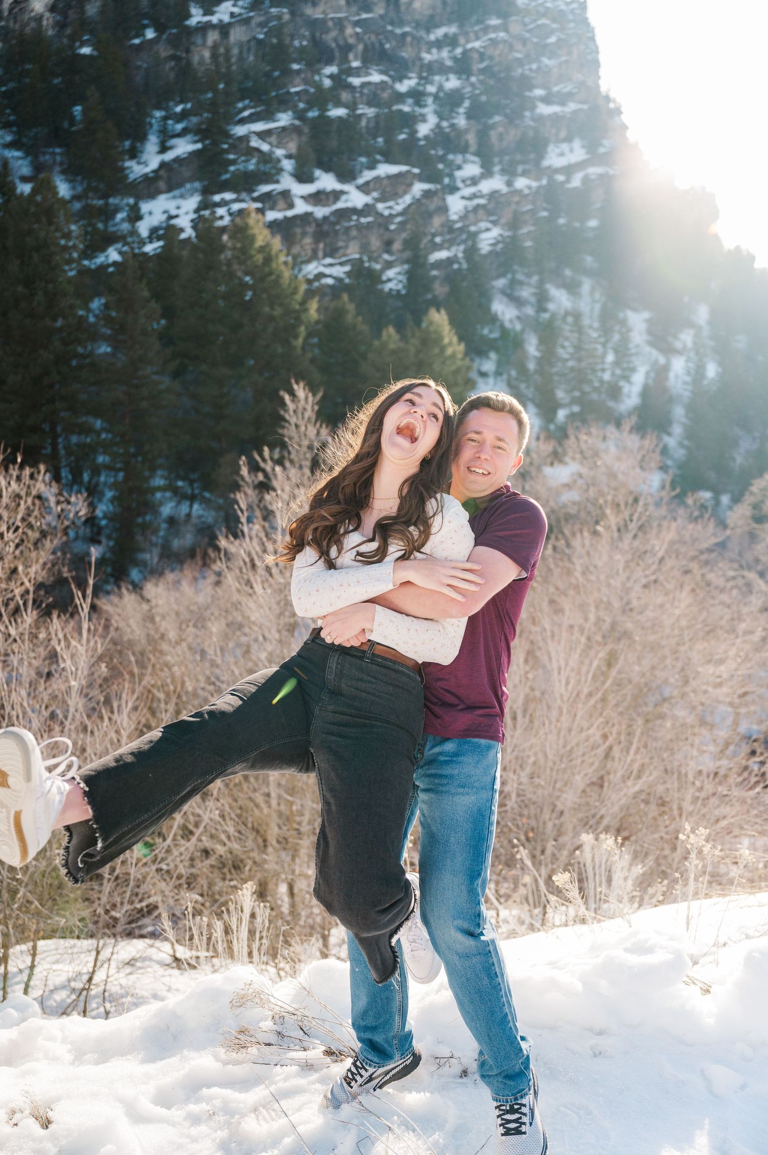 A couple laughing while sharing a playful moment in the snow with mountains visible behind.