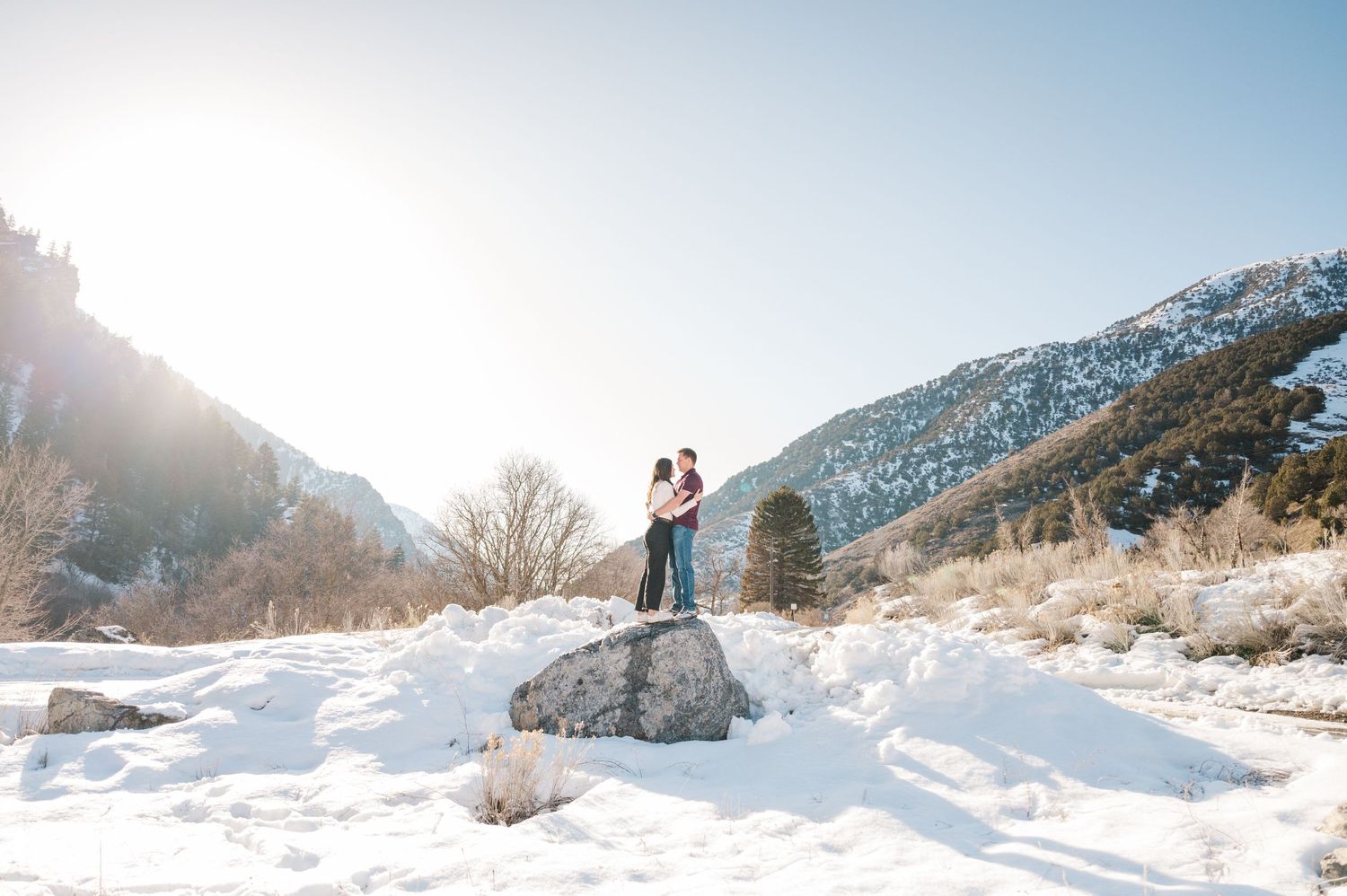 A couple stands atop a large rock surrounded by snow with mountains in the background.