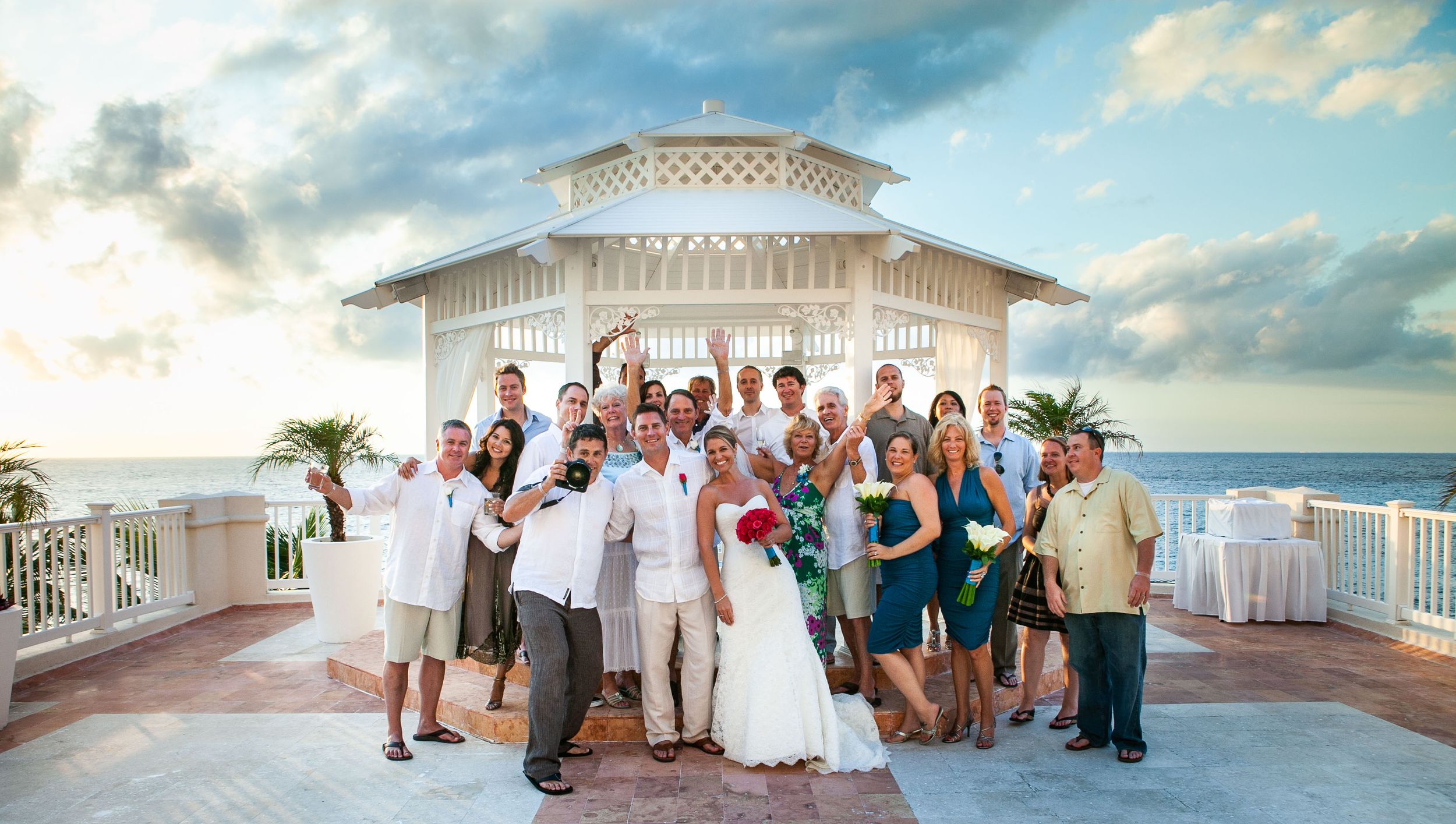 Wedding party poses for a group photo on a beachfront gazebo at sunset.