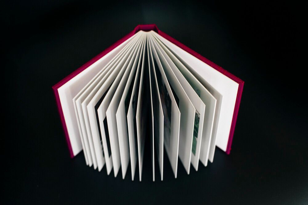 Red hardcover book fanned open to show layered white pages against dark background.