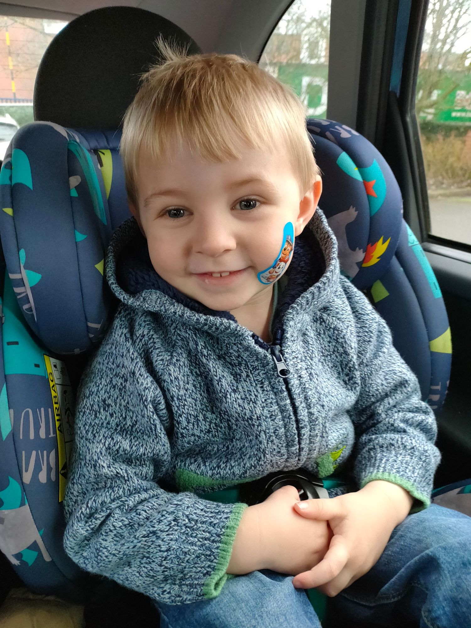Young child smiles while sitting in blue patterned car seat wearing gray fleece.