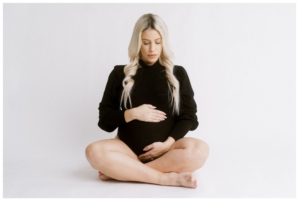 Maternity portrait of woman in black sweater sitting on white background.