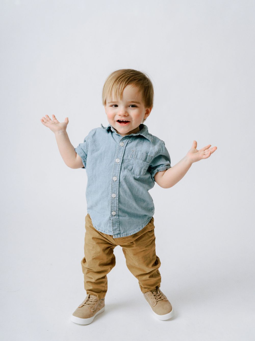 A toddler in denim and chambray shirt spreads arms wide with an excited expression.