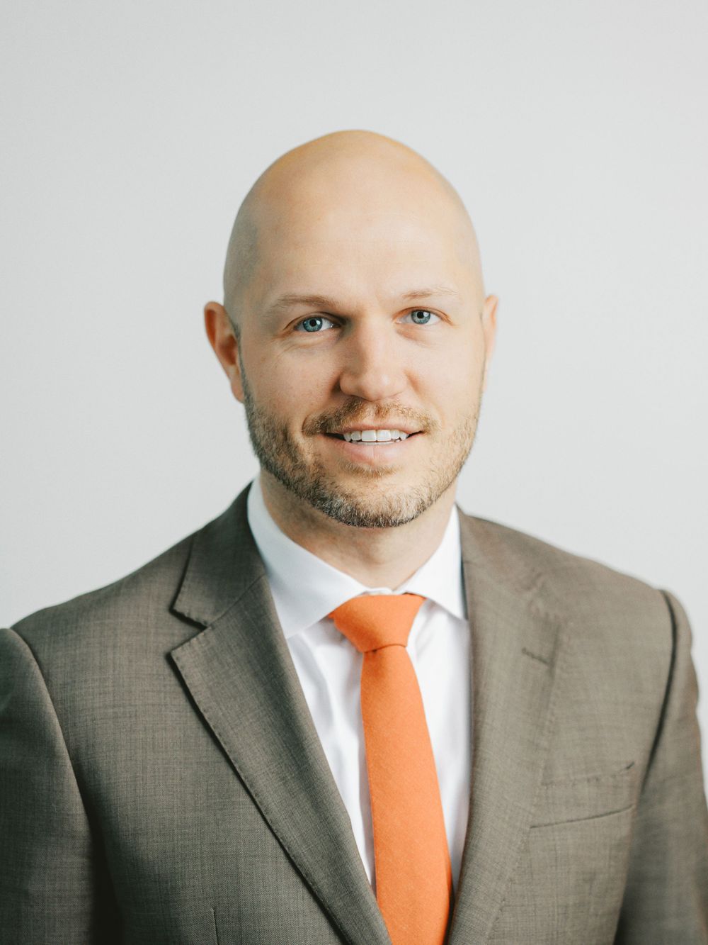 Professional headshot of person wearing brown suit with orange tie against light background.