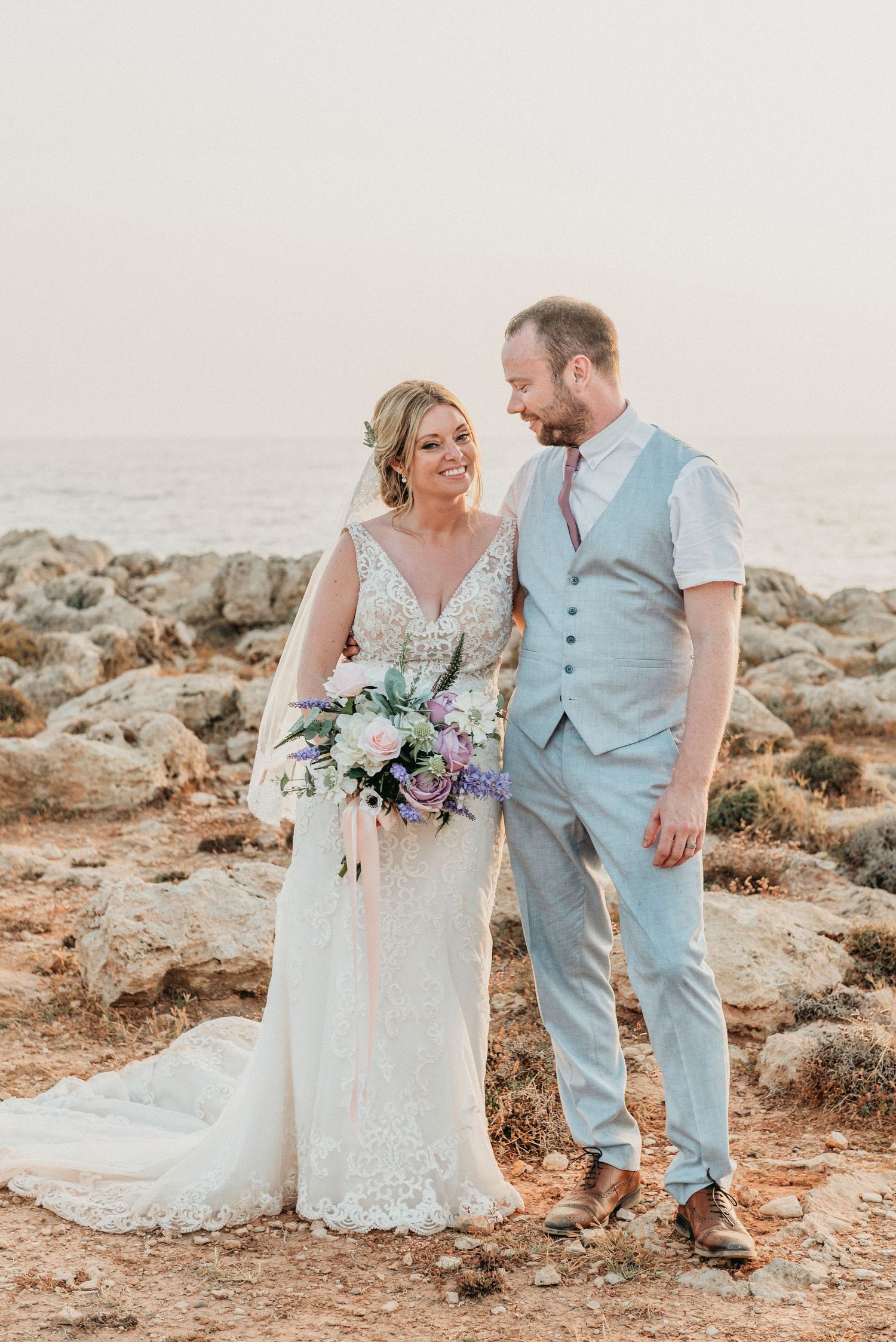 A scenic coastal wedding photo with a couple in formal attire holding a purple and white bouquet at sunset.