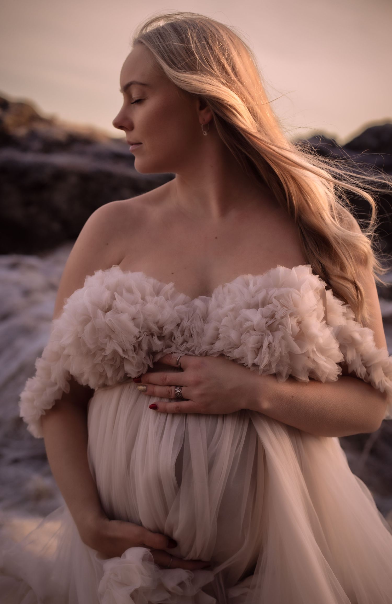 Pregnant woman in ruffled off-shoulder blush gown cradles belly among rocky coastal scenery at dusk.