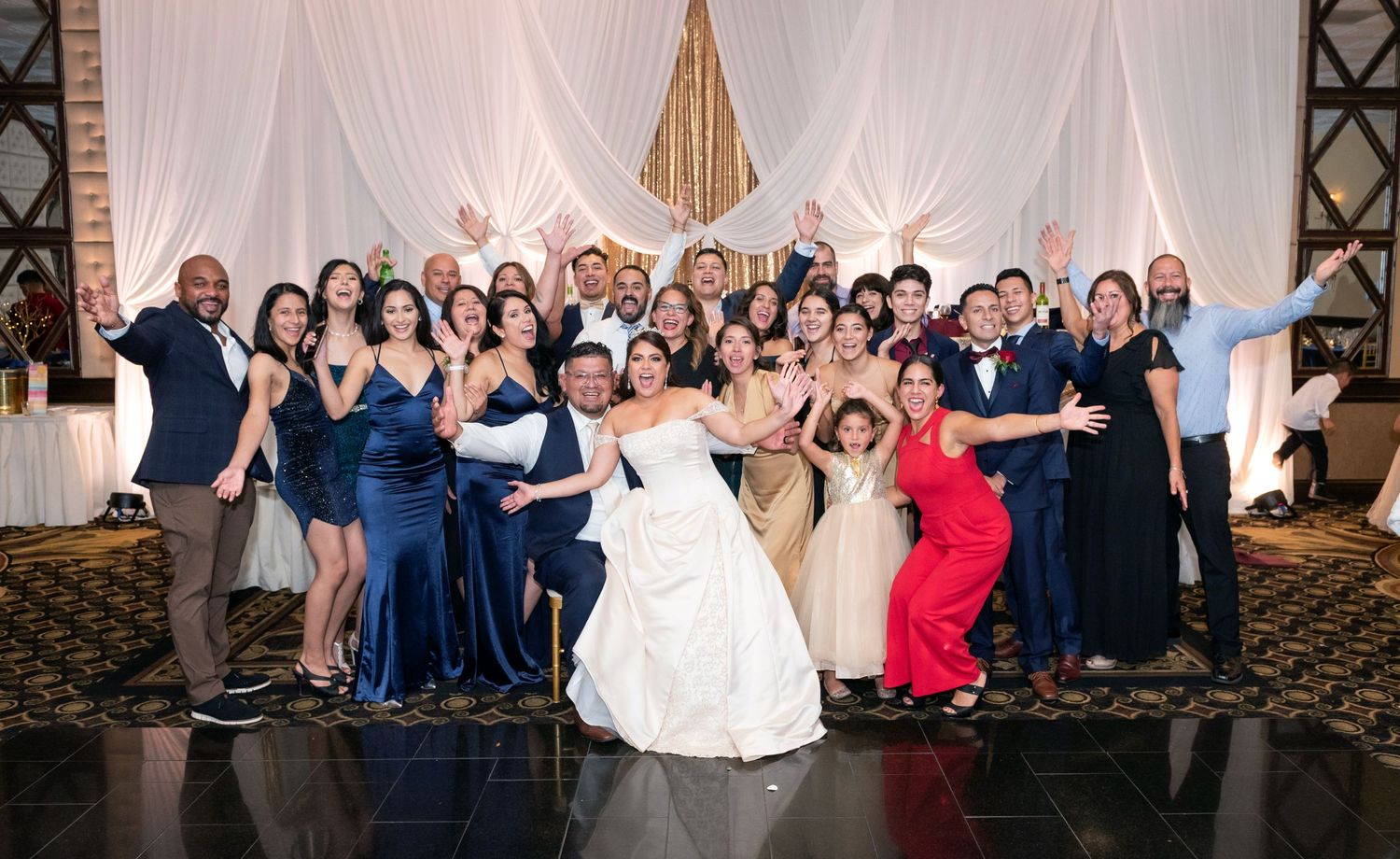 Large wedding party group photo in formal attire inside an elegant venue.