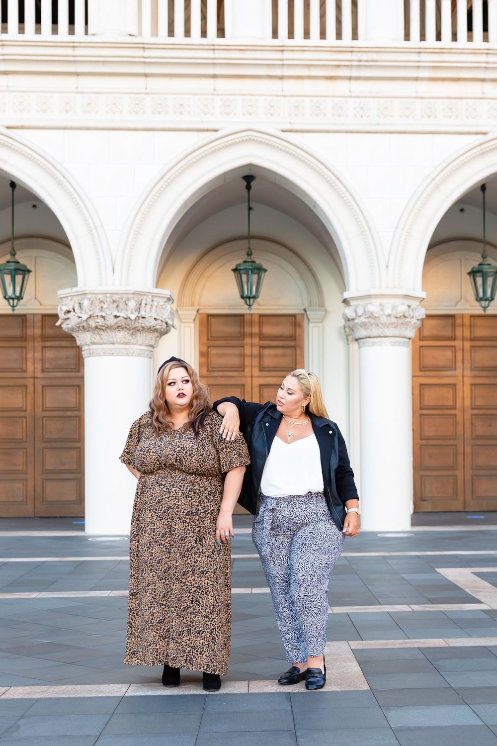 Two friends in stylish outfits stand beneath elegant white arches and hanging lanterns.