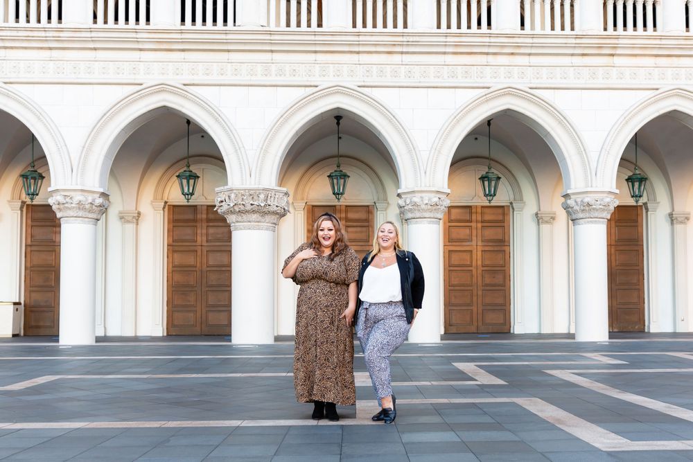 Two fashionable women pose together against ornate archways of a historic building.