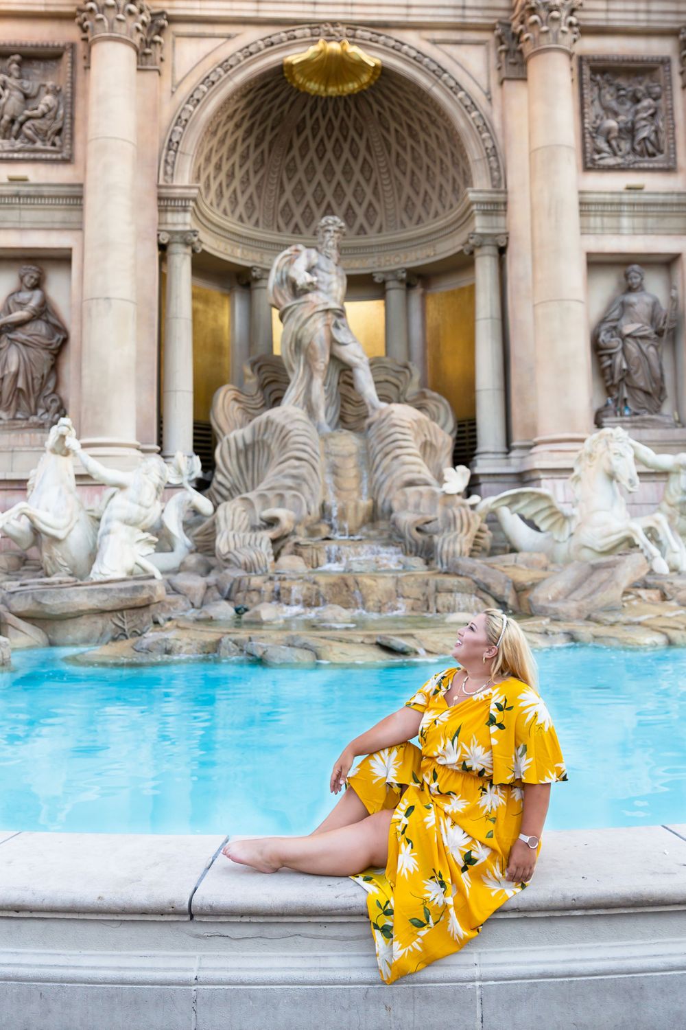 Tourist in vibrant yellow dress poses at the edge of the famous Trevi Fountain with baroque statues behind.