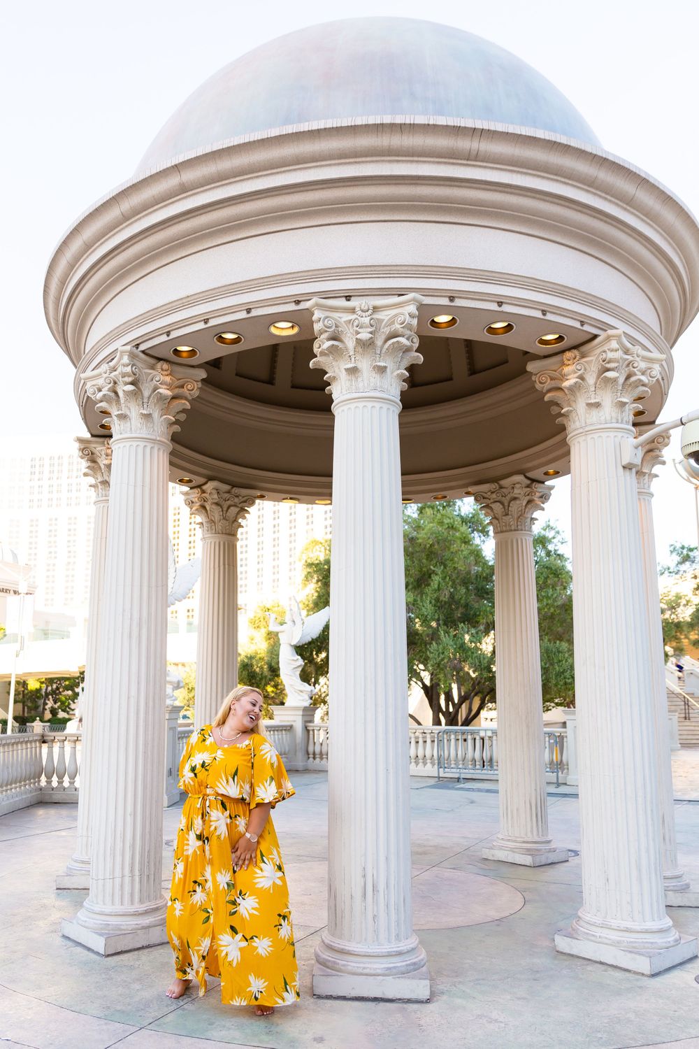 A yellow floral maxi dress flows elegantly under a classical white columned gazebo in an outdoor setting.