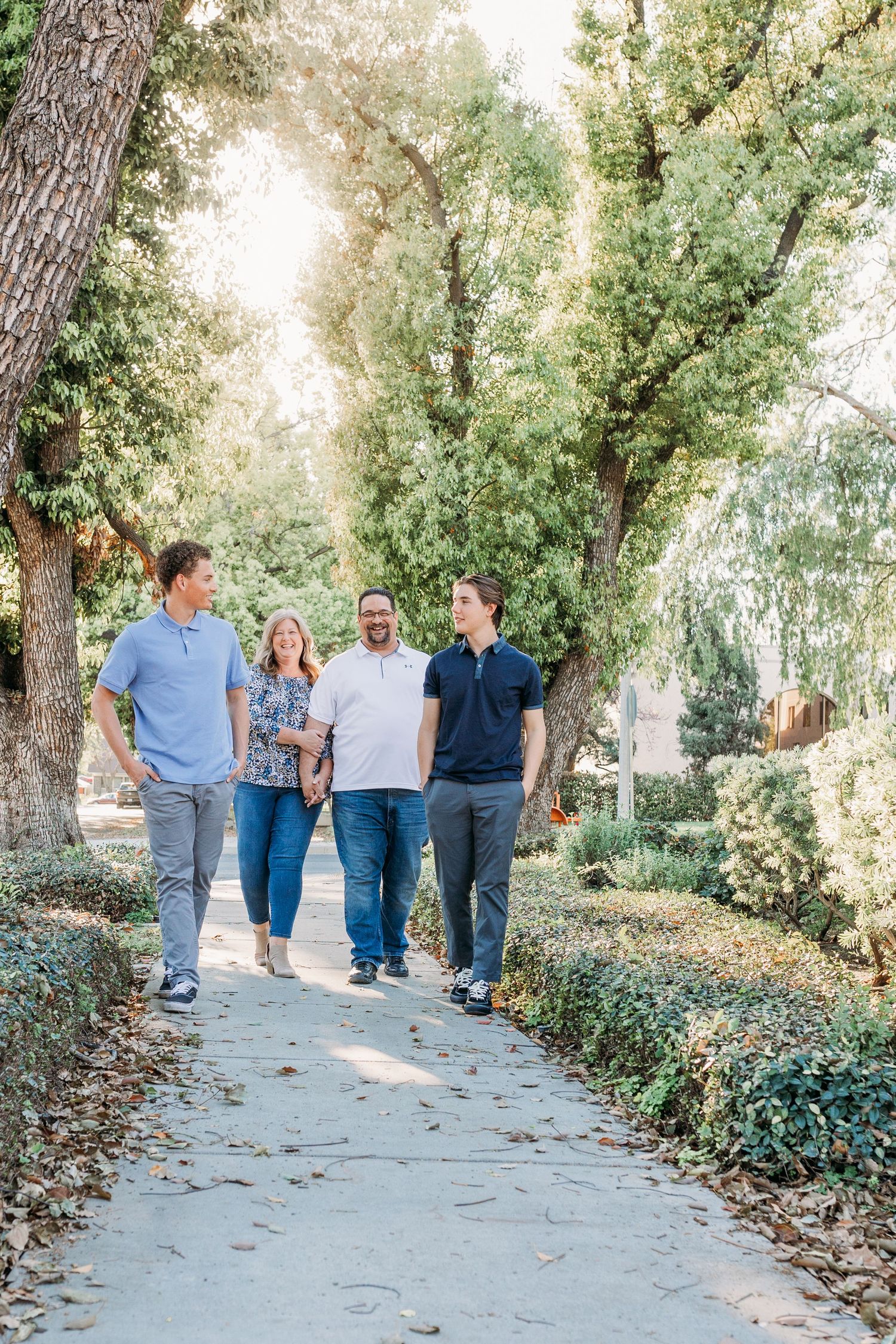 Family of 4 walking down a sidewalk surrounded by trees and sunshine.