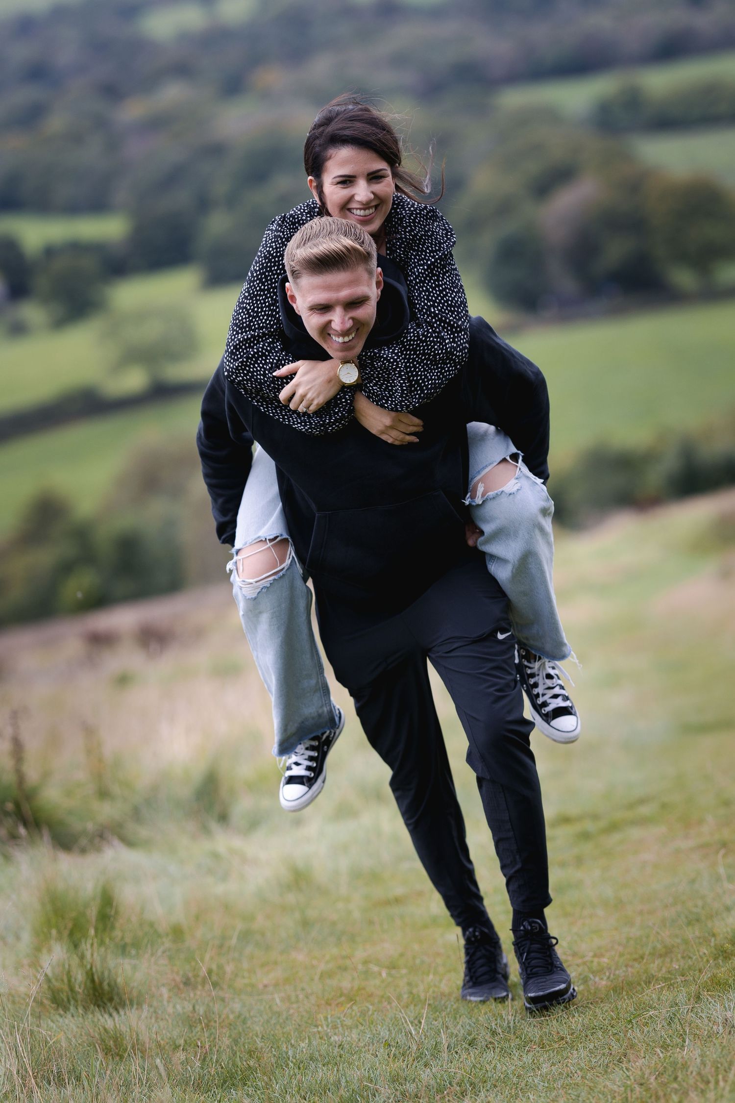 A couple shares a playful piggyback ride moment on a grassy hillside with scenic views.