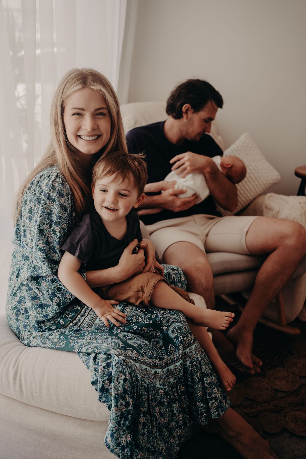 Family relaxes together on a couch, with someone wearing a floral dress holding a child while another feeds a baby.
