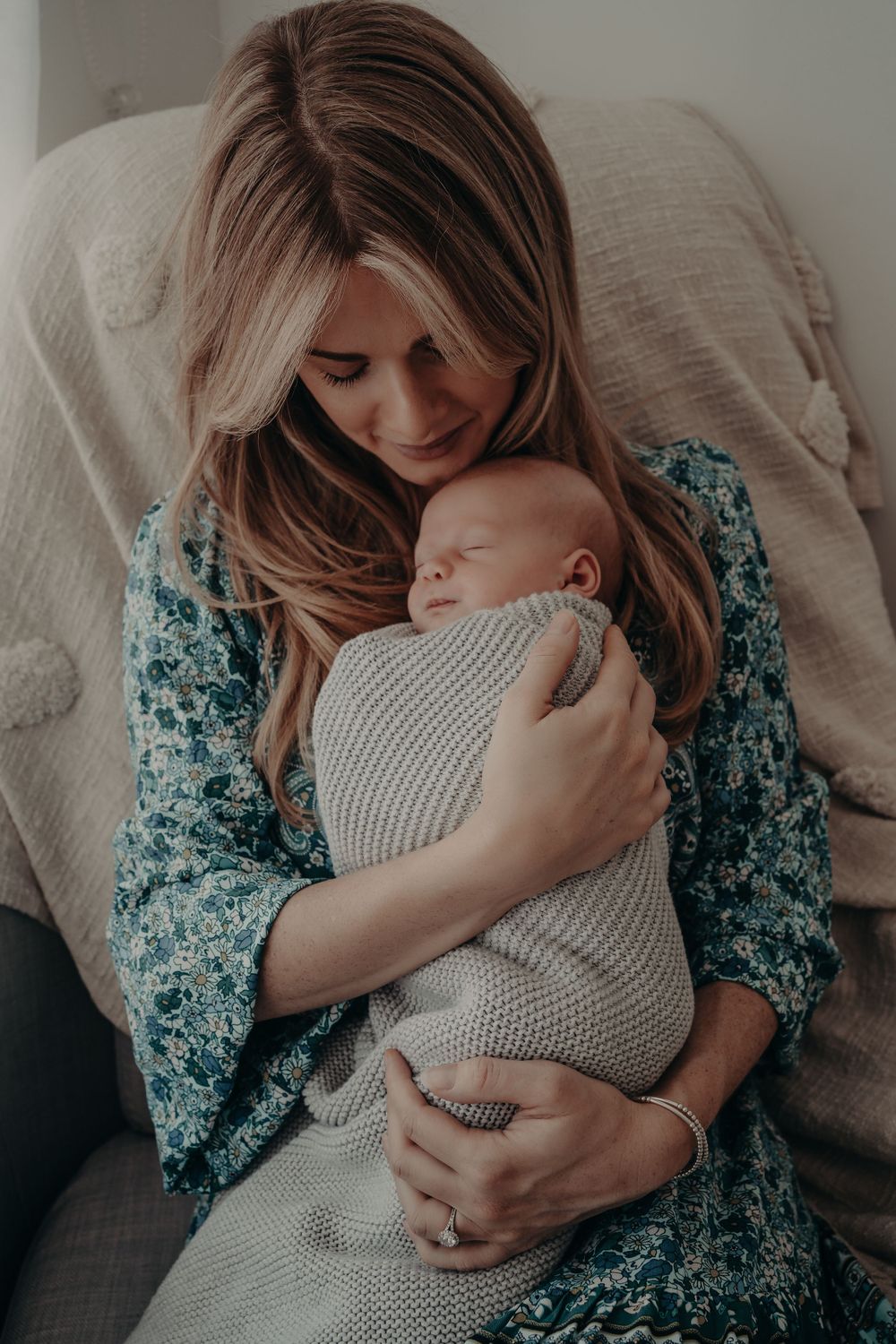 Someone in a floral dress tenderly holds a sleeping baby wrapped in a knit blanket.