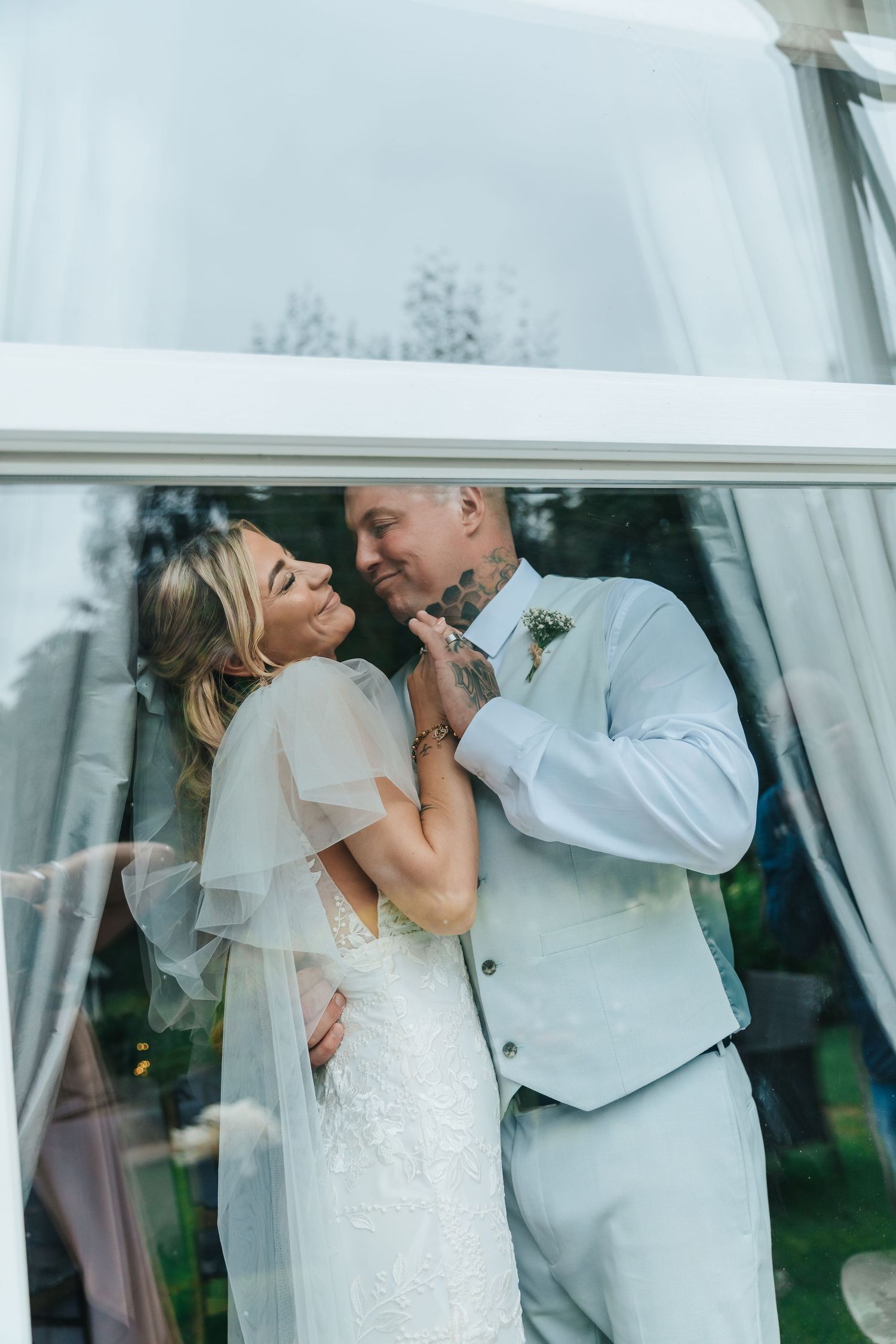 A romantic wedding moment between newlyweds in white attire behind sheer curtains.