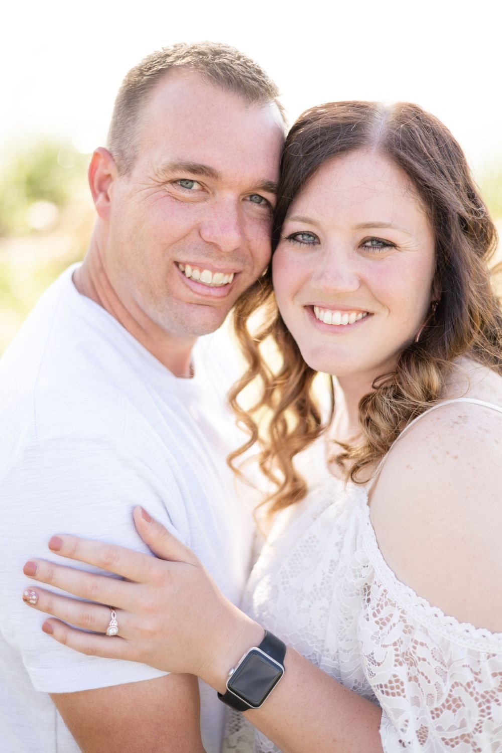 A close-up portrait of a couple in white clothing smiling outdoors in bright sunlight.